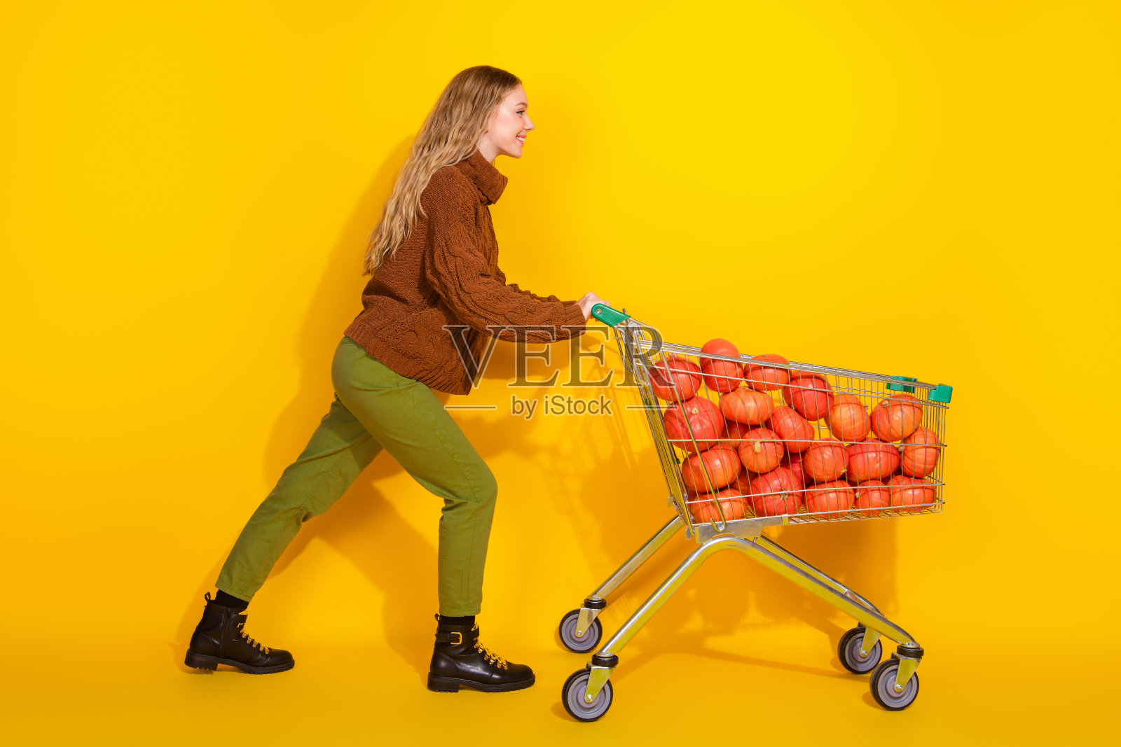 漂亮年轻女性推着购物车，车里装着南瓜，背景为黄色，空旷的空间。照片摄影图片
