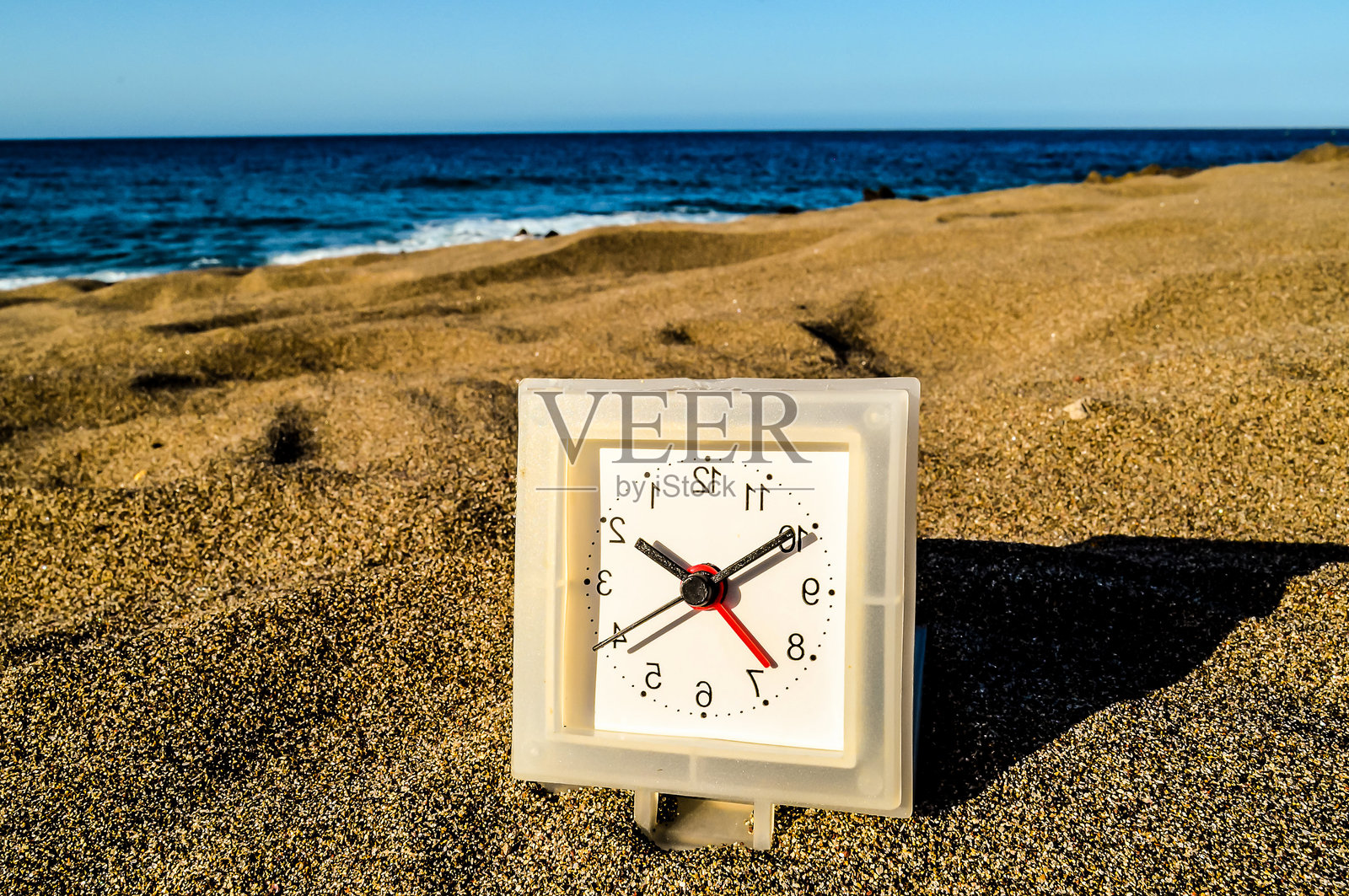 一个小白色时钟静静地放在海边的沙滩上。照片摄影图片