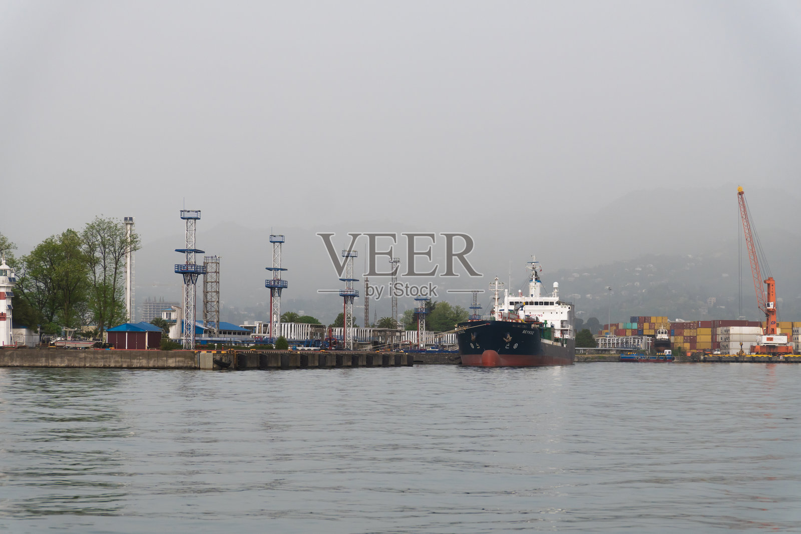 货船在雾蒙蒙的日子里驶入繁忙的港口，经过灯塔和工业设备。起重机、集装箱和雾气构成了热闹的场景。照片摄影图片