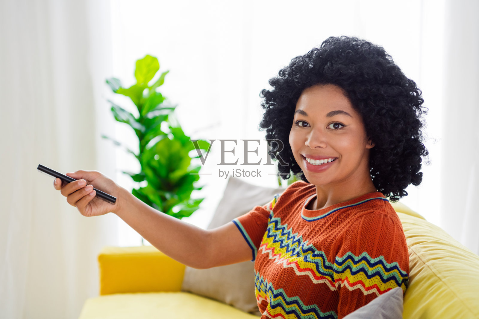 年轻女性在现代公寓室内穿着T恤，手持电视遥控器，展现出周末的轻松氛围。照片摄影图片