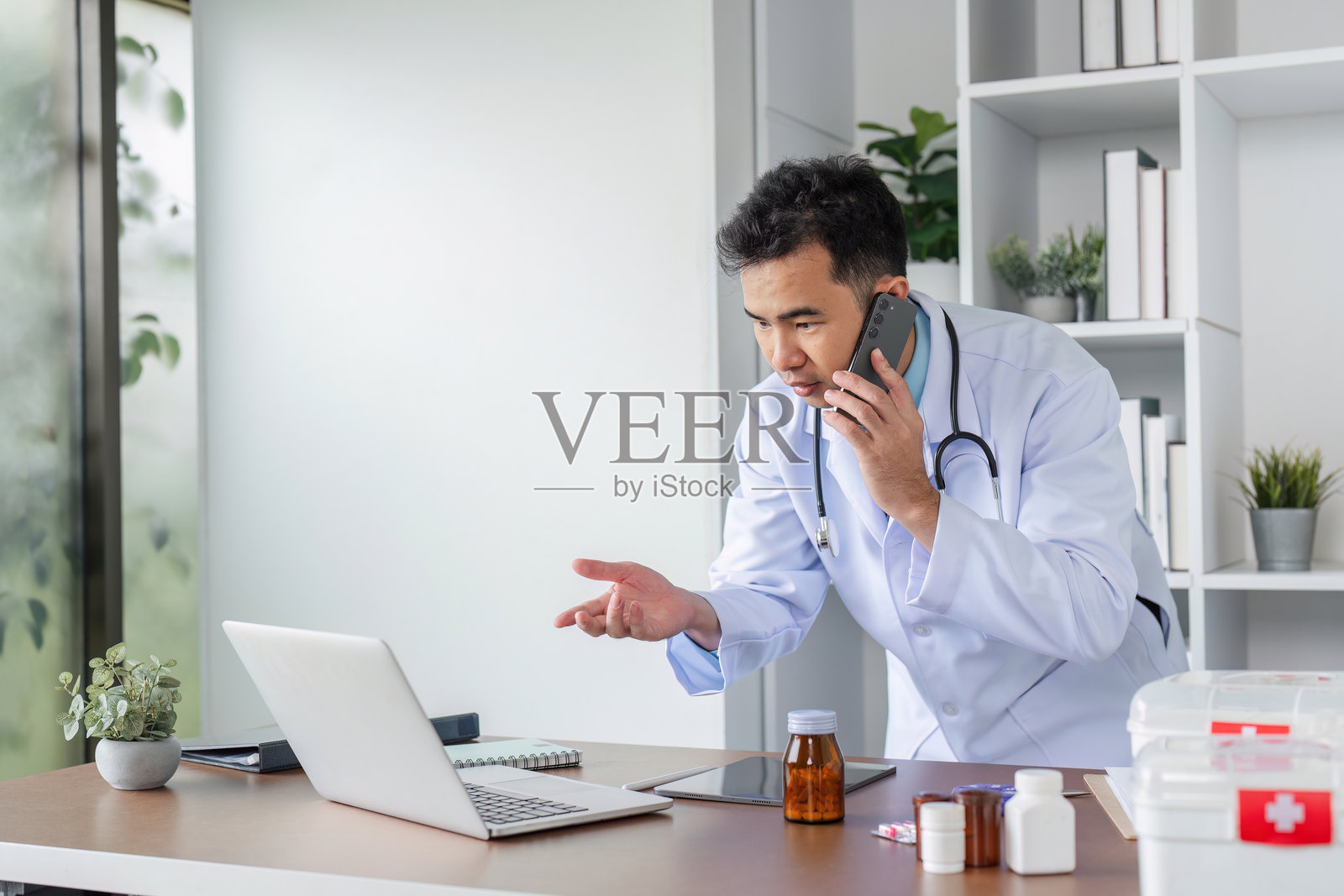 自信的男性医生穿着白大褂，在现代医疗办公室环境中通过电话讨论病人护理。照片摄影图片