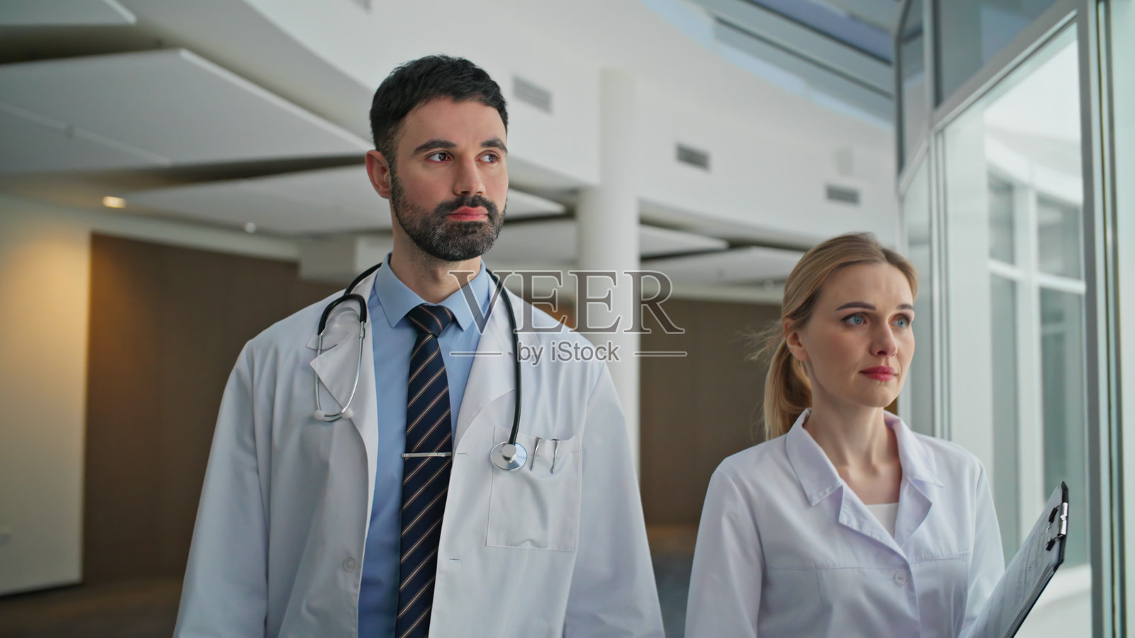 穿着白大褂的临床工作人员在走廊中走动。特写镜头下，两位医生看向镜头。照片摄影图片
