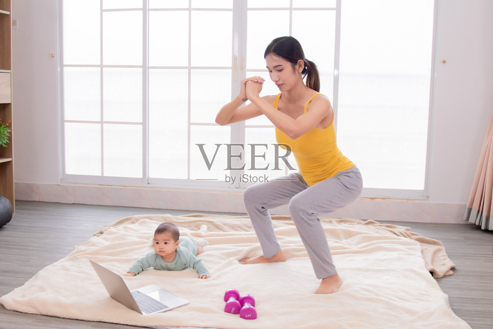 年轻的亚洲母亲在笔记本电脑上观看视频并做深蹲锻炼，旁边的新生女儿好奇地看着她，体现了关注身体健康的理念。照片摄影图片