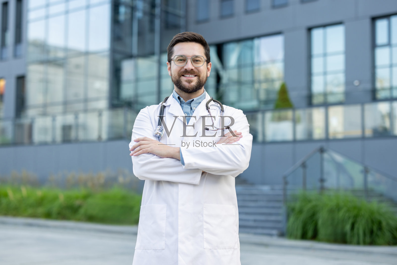 一位年轻成功、面带微笑的男性医生站在医院建筑外，穿着白大褂，肩上挂着听诊器，双臂交叉在胸前，正看着镜头。照片摄影图片