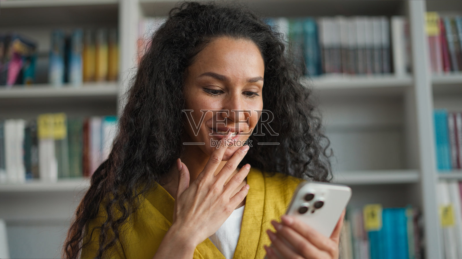 兴奋的拉丁裔白人西班牙裔女性手持手机，面带惊喜的微笑，情绪激动，正在查看高中或大学的考试成绩，身处图书馆，作为学生的她对社交媒体和学习感到非常高兴。照片摄影图片