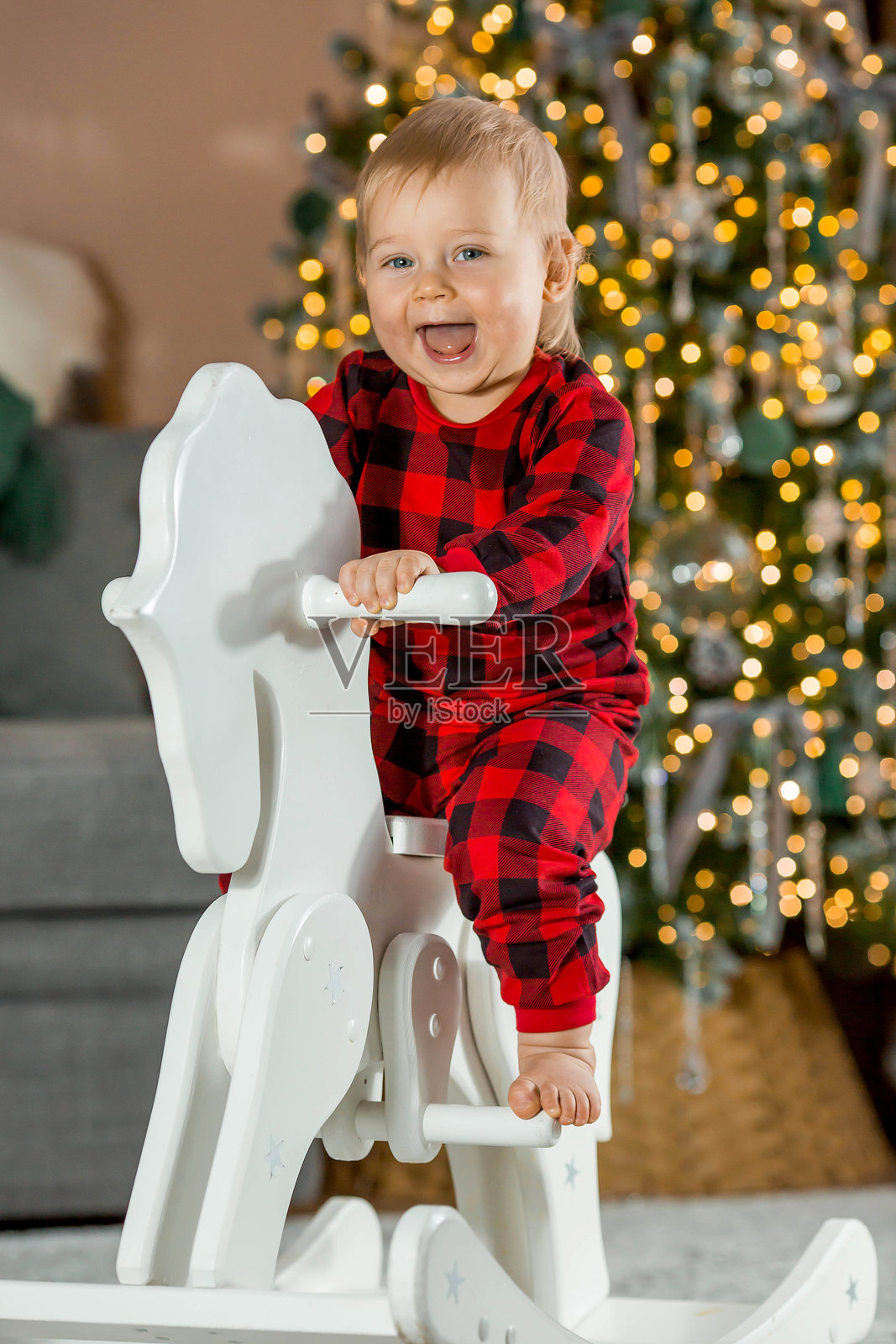 家庭。一个美丽的1岁金发小男孩坐在木马旁，背景是圣诞树。庆祝圣诞节和新年的概念。照片摄影图片