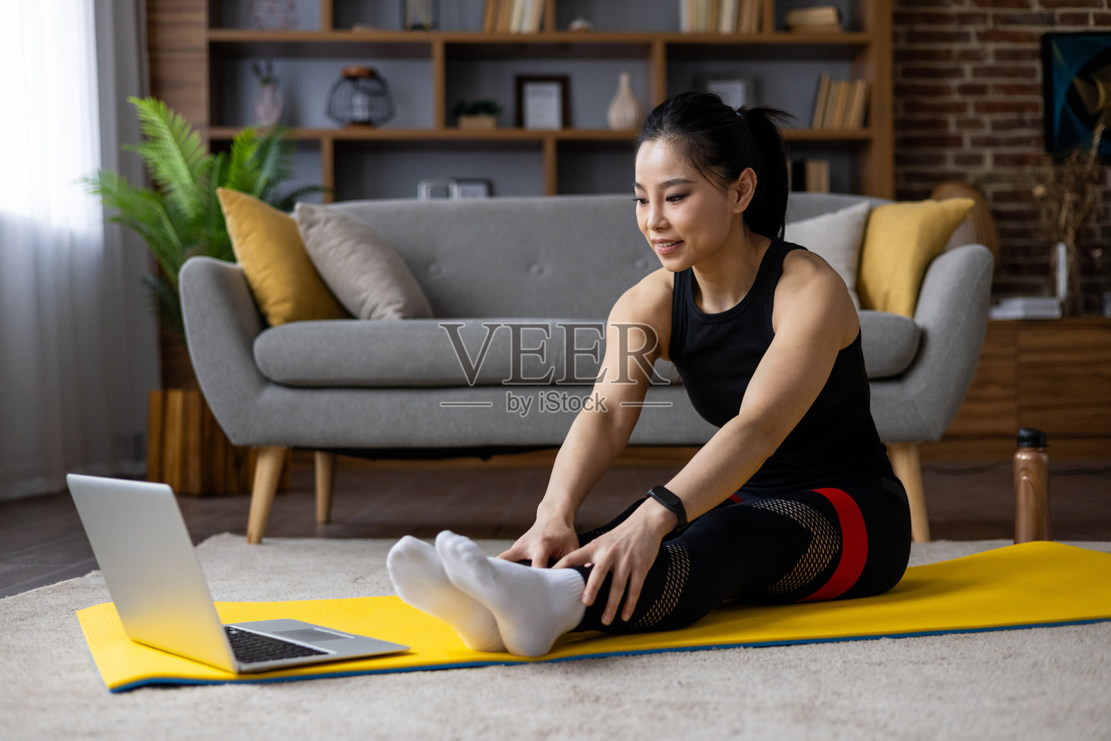 亚洲女性在家中黄色瑜伽垫上做拉伸，跟随笔记本电脑上的在线健身课程。舒适的客厅环境，现代装饰和自然光线。照片摄影图片