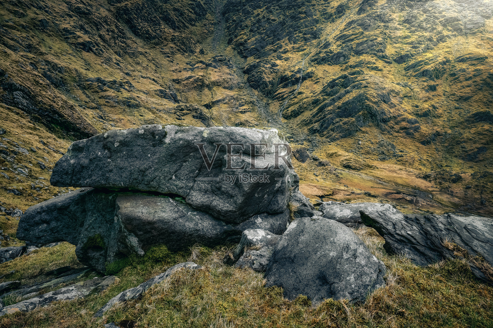 一块大石头坐落在草坡上，周围是崎岖的山谷。风景中有干燥的草地和岩石地形，天空布满云彩。照片摄影图片