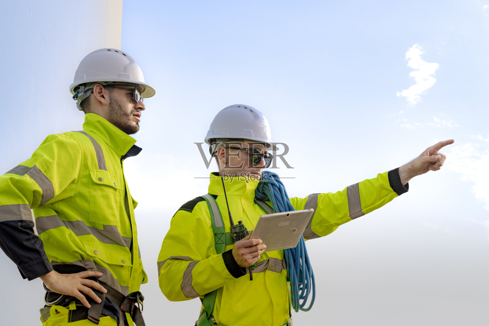两名穿着高可视性服装和安全帽的工程师在风能现场观察风力涡轮机，并通过无线电进行沟通。该图像突出了团队合作、安全和可再生能源生产。照片摄影图片