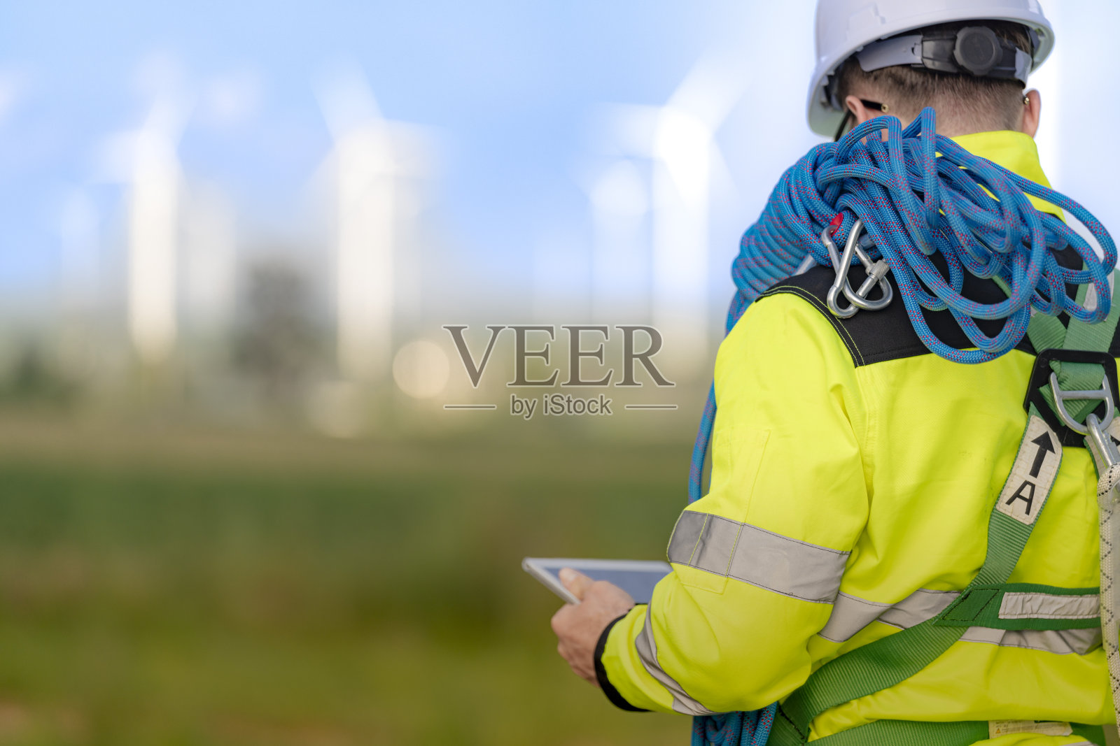 两名穿着高可视性夹克和安全帽的工程师正在检查风电场。一名工程师手持移动设备，另一名则携带攀爬用的绳索，背景中可见风力涡轮机。照片摄影图片