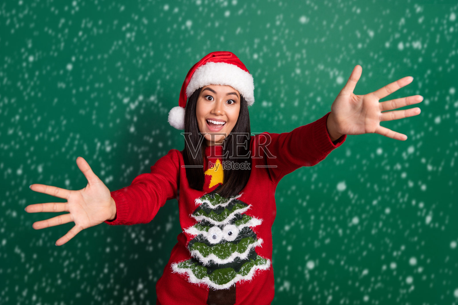 年轻可爱的韩国女孩穿着红色旧毛衣，满意而平静，想要拥抱你，背景是绿色的树饰。照片摄影图片