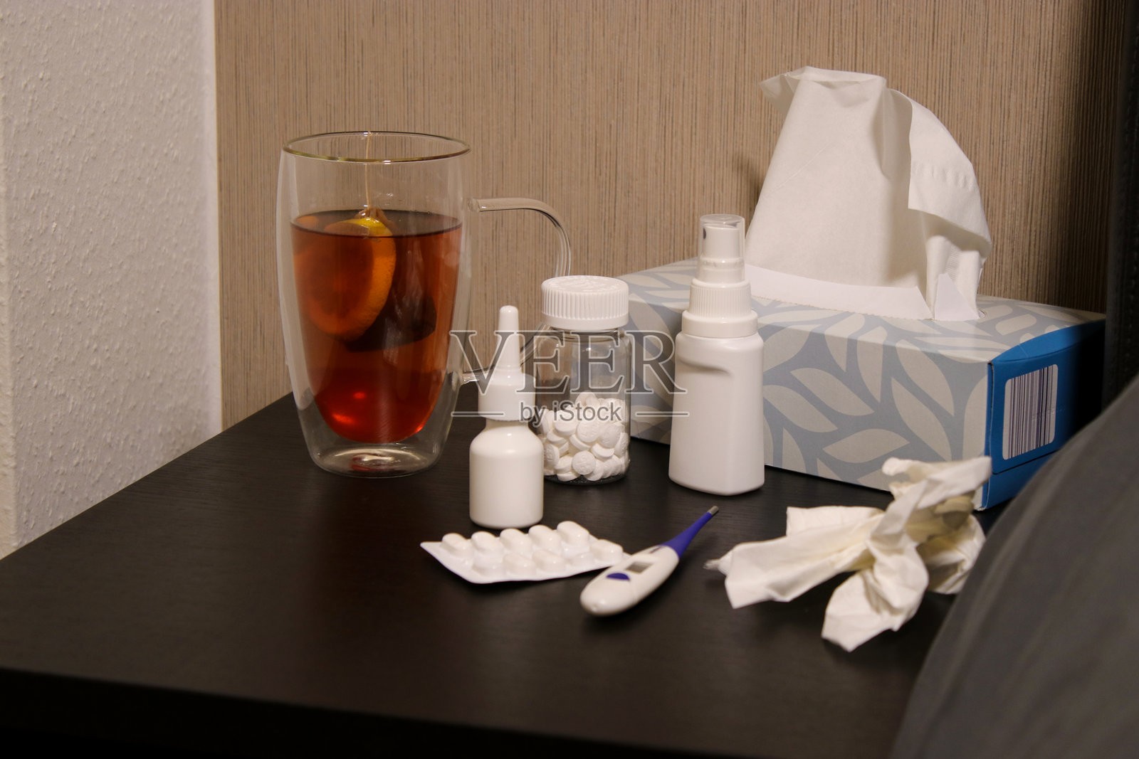 床头柜上放着热茶、柠檬、药丸、鼻喷雾、喉喷雾和温度计，体现生病在家的概念。照片摄影图片