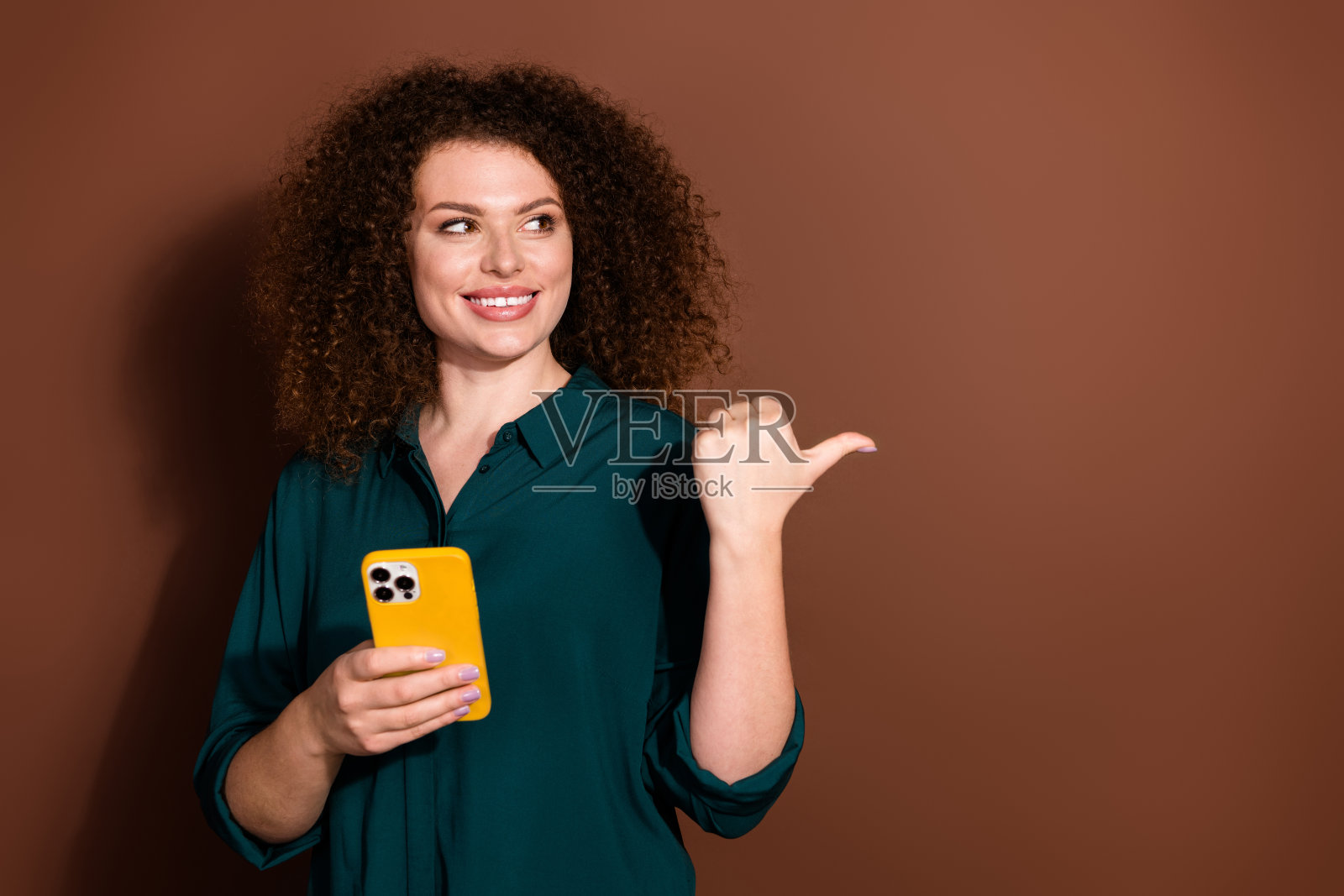 迷人积极的女性穿着绿色衬衫，竖起大拇指，手持现代设备，背景为空白的棕色。照片摄影图片