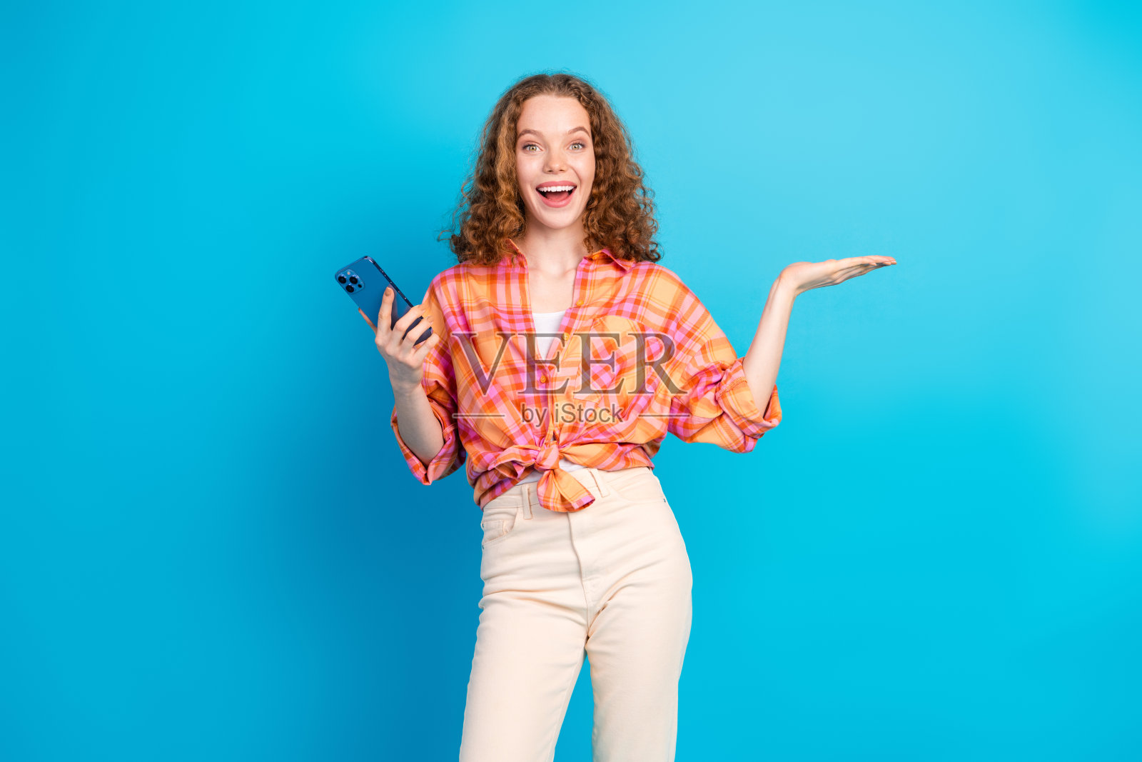 快乐的年轻女性在充满活力的蓝色背景前展示概念，同时手持智能手机。照片摄影图片