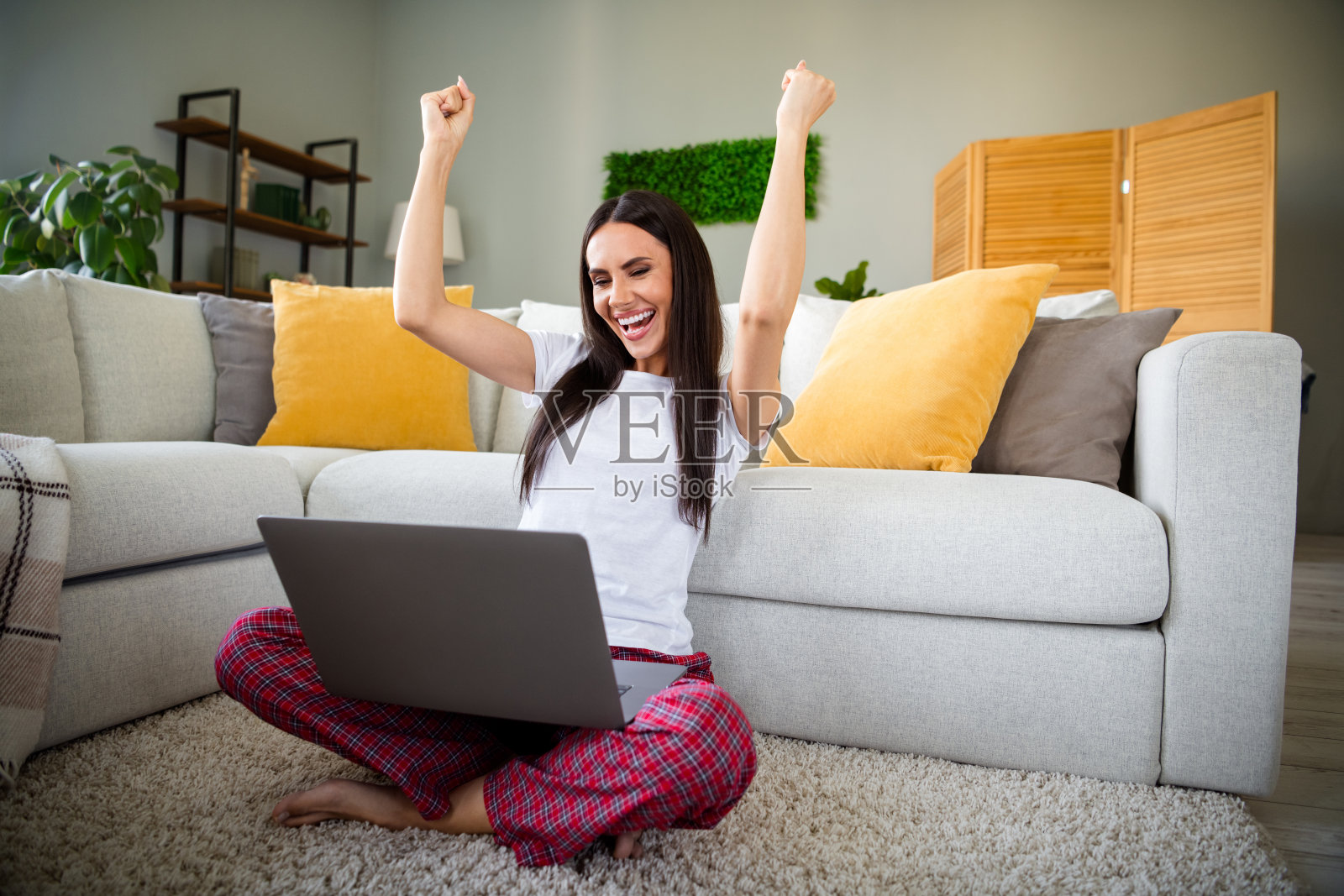 年轻漂亮的女士坐在地板上，举起双拳，手持笔记本电脑，享受周末时光，现代公寓室内环境。照片摄影图片