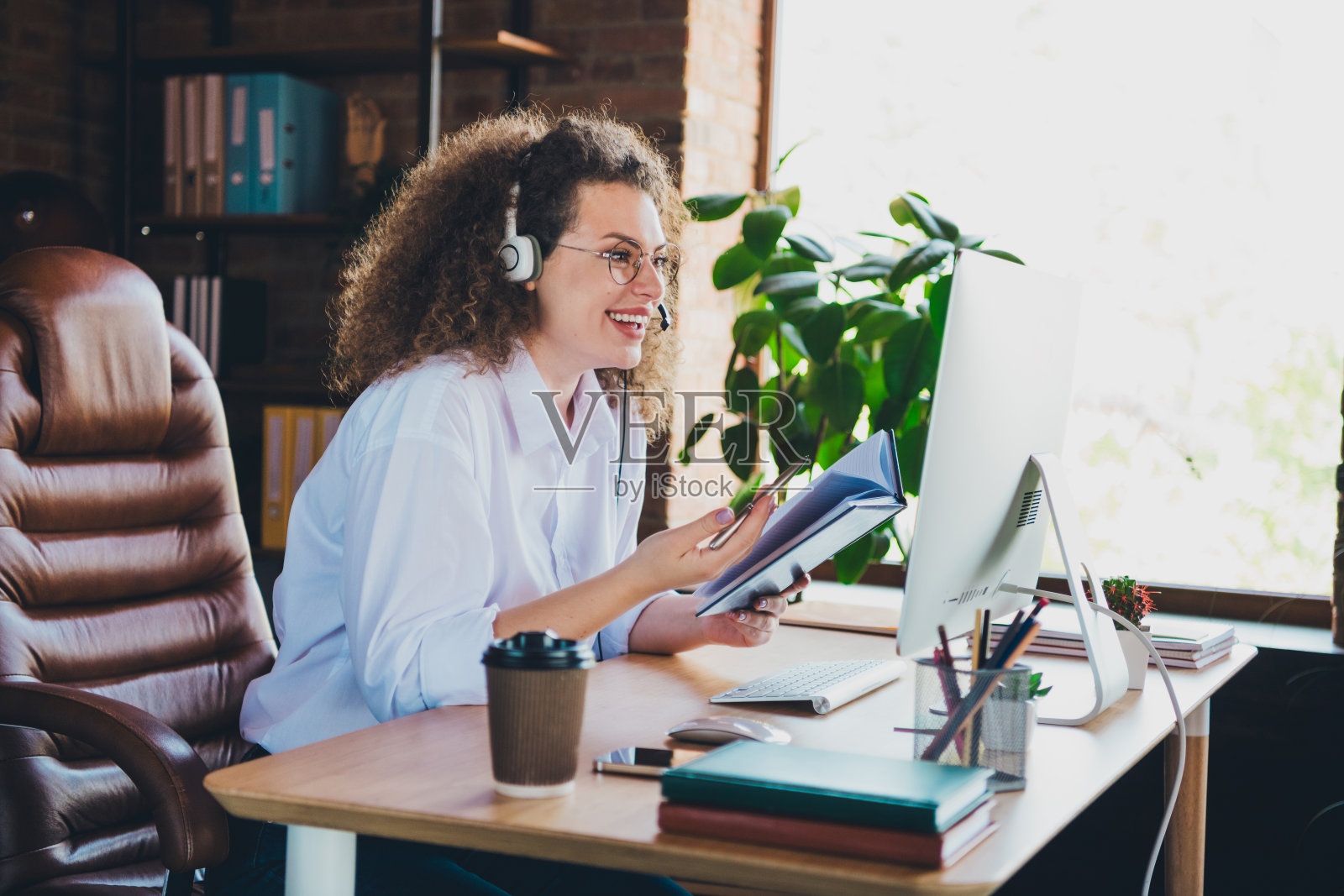 漂亮的卷发年轻女孩在呼叫中心进行视频通话，穿着正式的商务服装，作为顾问在成功的共享办公环境中工作，光线明亮。照片摄影图片