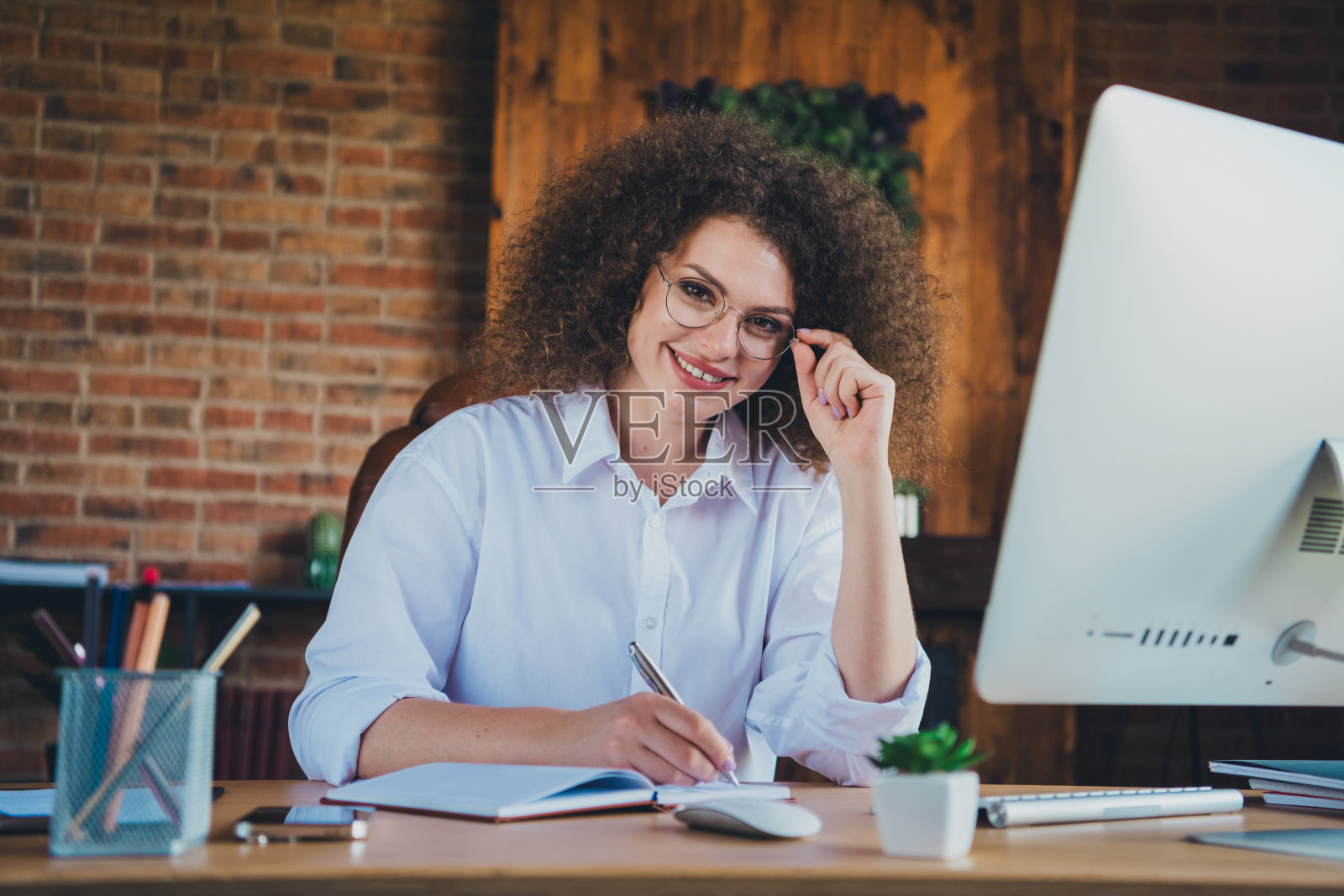 穿着正式服装的迷人卷发年轻女性在现代办公室的咨询室内写笔记本照片摄影图片