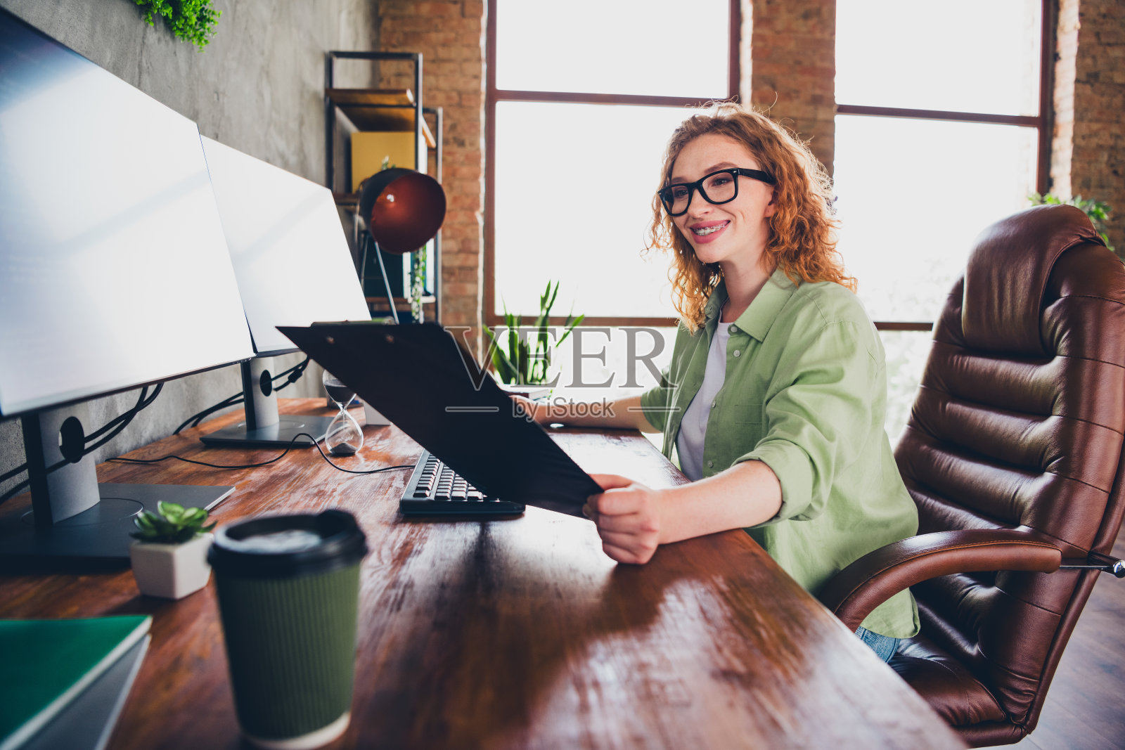 自信积极的女招聘者穿着衬衫，戴着眼镜，在现代设备上打字，阅读文件，坐在室内工作场所的工作站中。照片摄影图片