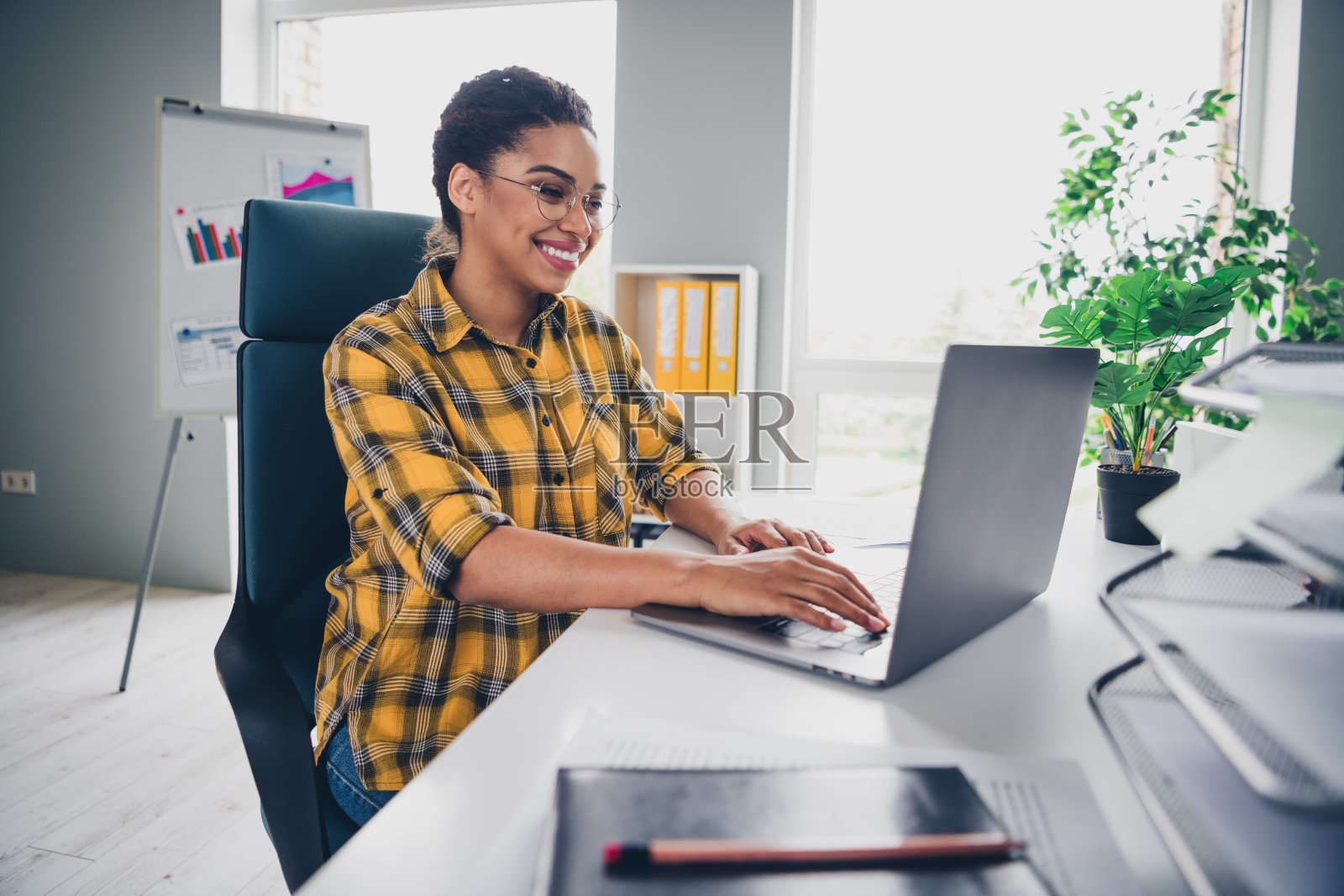 穿着黄色格子衬衫的迷人年轻女性在现代办公室阁楼内的舒适工作空间使用笔记本电脑。照片摄影图片