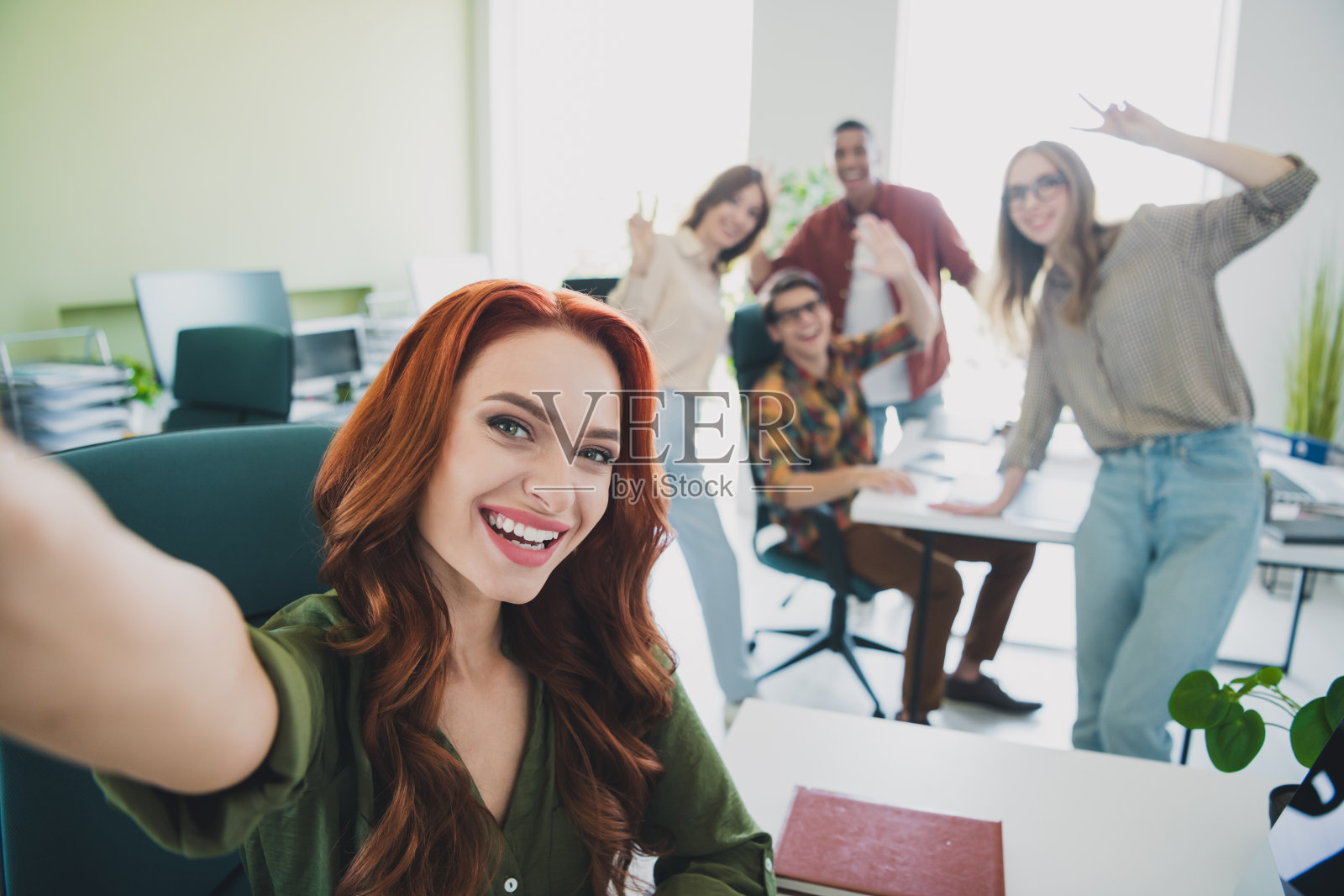 一群年轻的企业员工在办公室商务中心的工作站拍摄自拍，做出V字手势。照片摄影图片