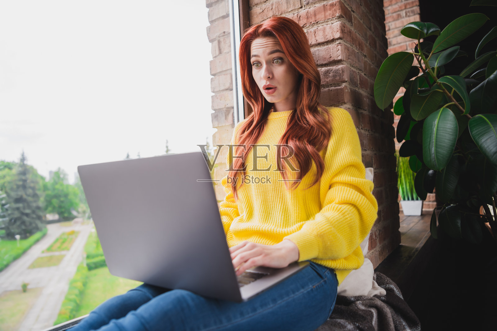 漂亮年轻女性在公寓室内穿着黄色毛衣，张嘴盯着笔记本电脑。照片摄影图片