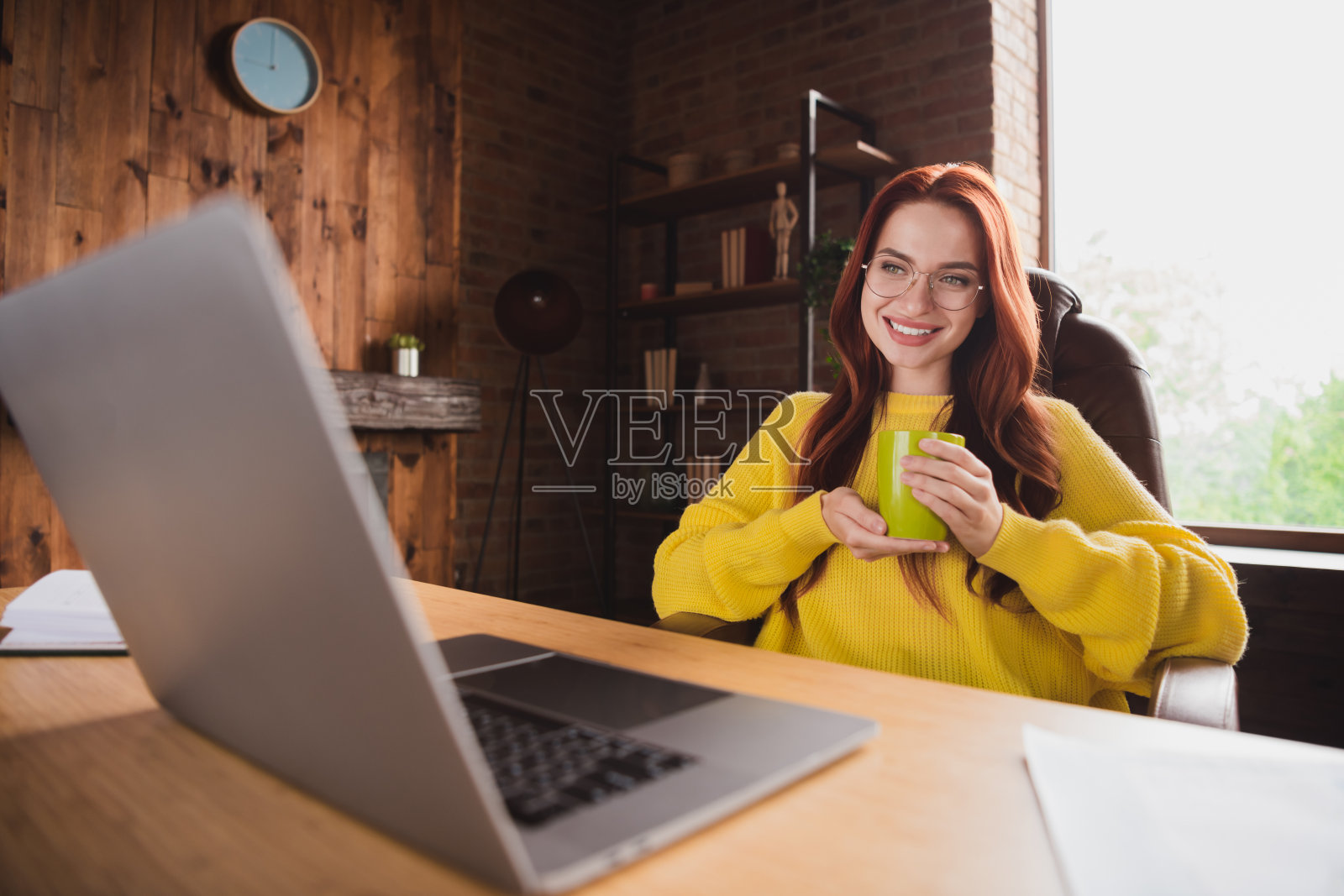 年轻女孩在家办公室的桌子上拿着新鲜咖啡和笔记本电脑，背景是阁楼式的室内环境。照片摄影图片