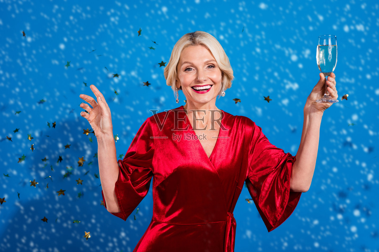 兴奋的时尚退休女士穿着闪亮的红色服装，举起酒杯享受圣诞派对，背景为蓝色。照片摄影图片