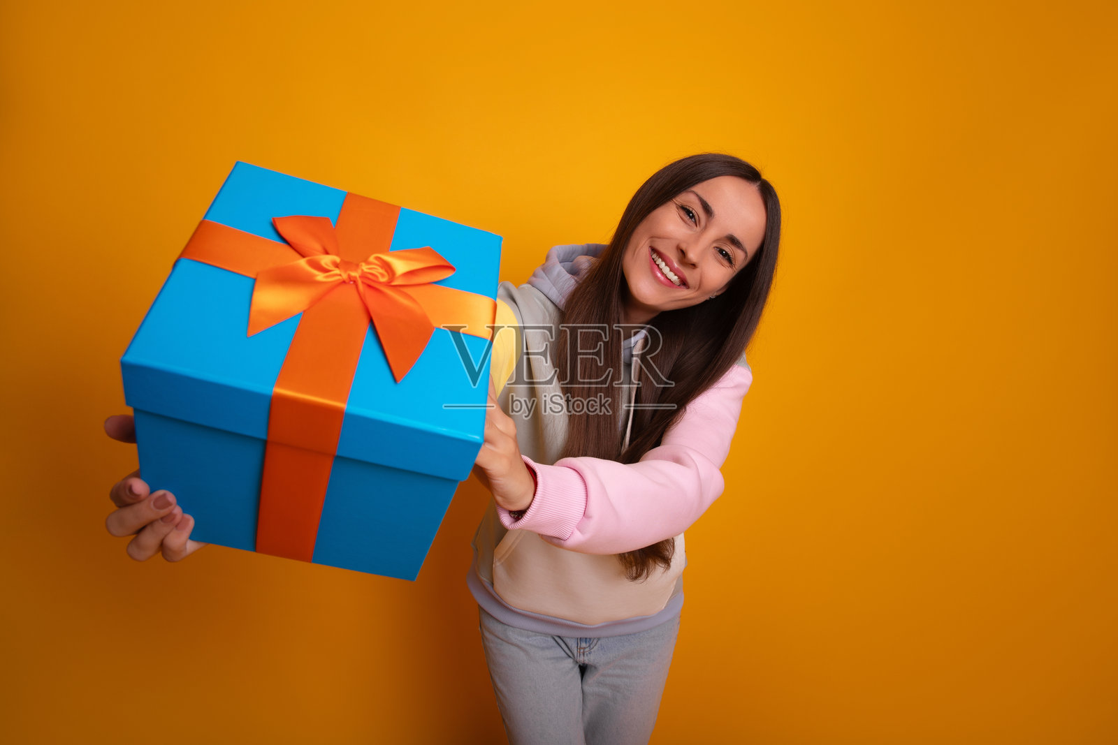 一位快乐的年轻女性微笑着向镜头展示一个大蓝色礼盒，上面系着橙色丝带。她站在鲜艳的黄色背景前，散发着快乐和慷慨的气息。照片摄影图片