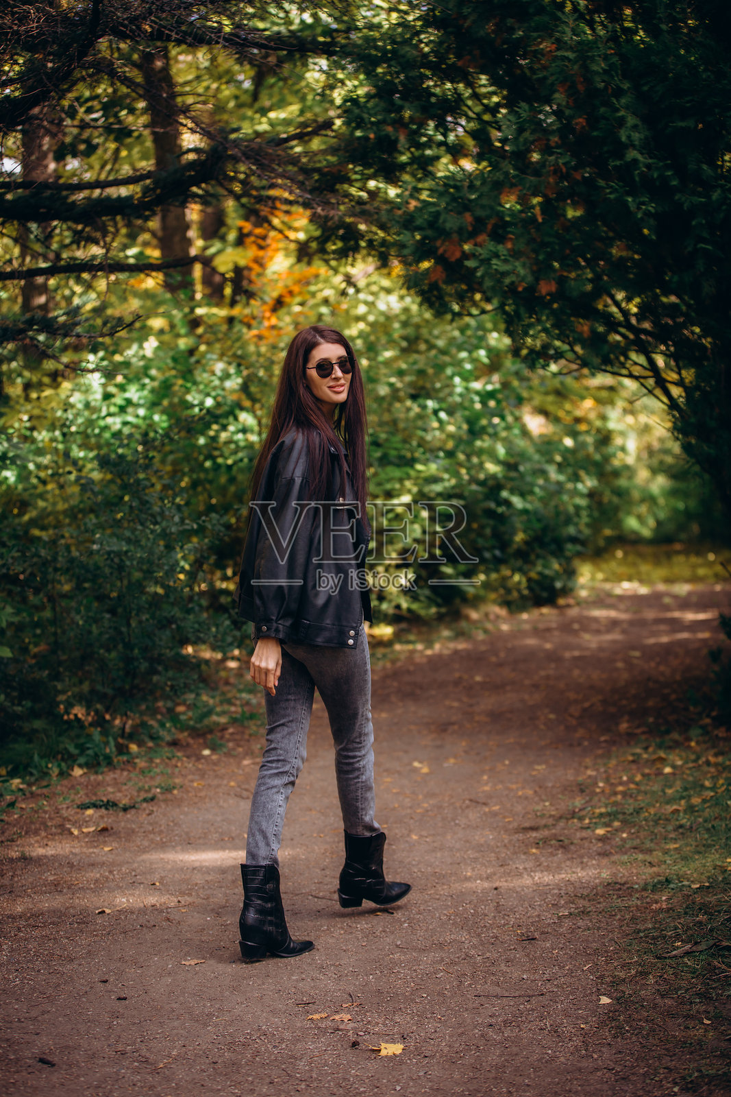 一位成熟的女性，身穿皮夹克和牛仔裤的美丽棕发女子，在秋天的公园里散步。照片摄影图片