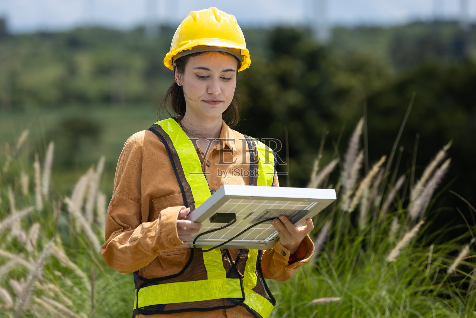 女性工程师在生态太阳能电池板厂环境中工作。替代创新能源。女性手持太阳能电池板，背景是风力涡轮机。照片摄影图片