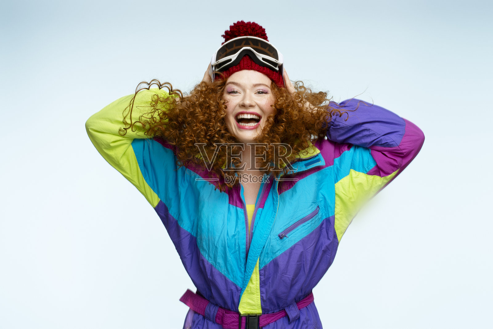 红发女性穿着色彩斑斓的滑雪服和护目镜，微笑着双手放在头上照片摄影图片