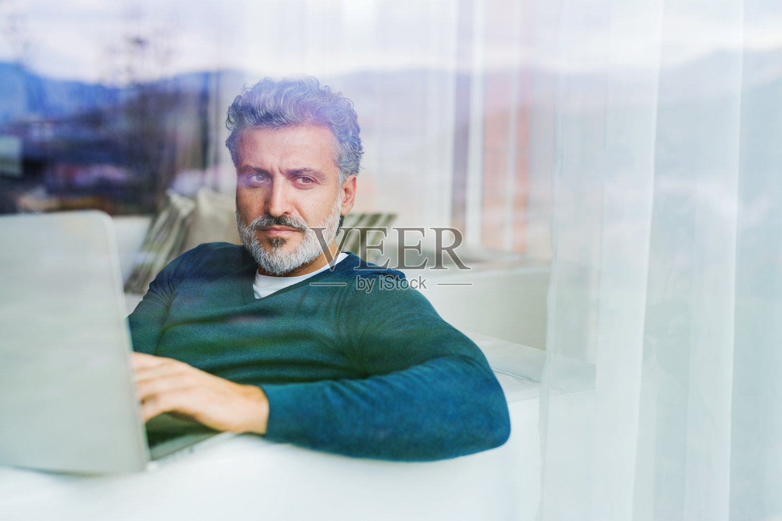 坐在沙发上用笔记本电脑工作的帅气男士肖像，透过窗户拍摄。照片摄影图片