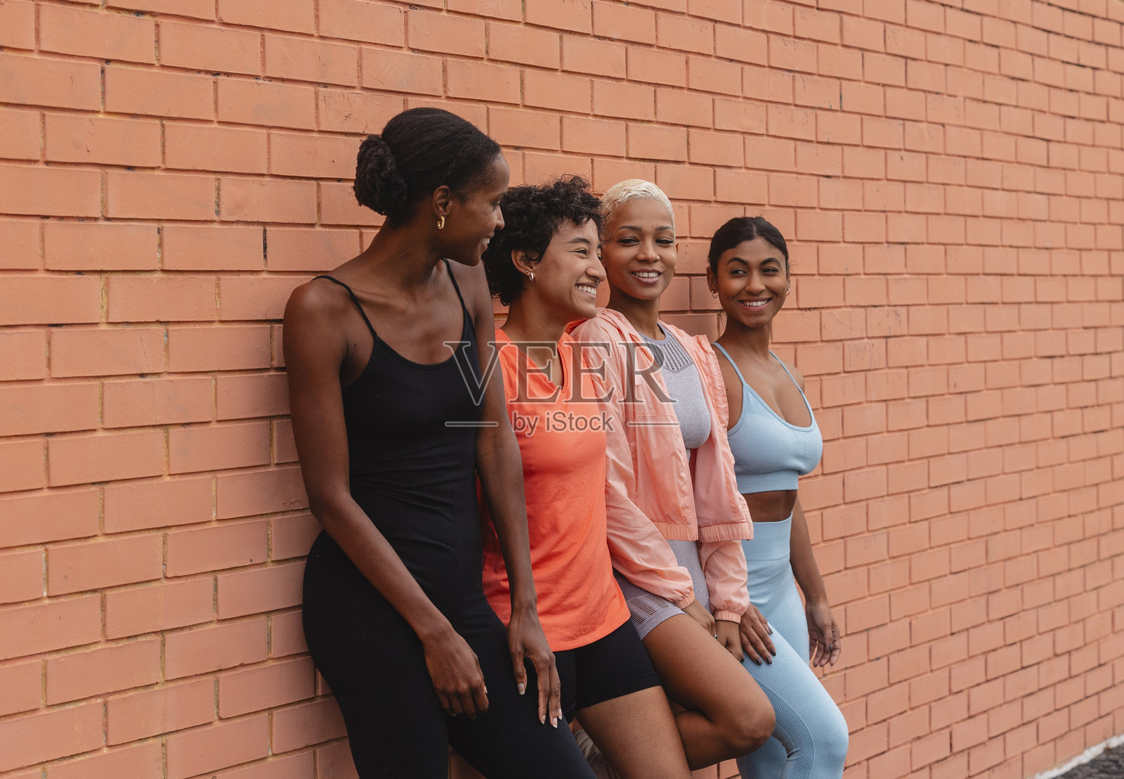 四位女运动员靠在砖墙上微笑放松，训练后休息。照片摄影图片