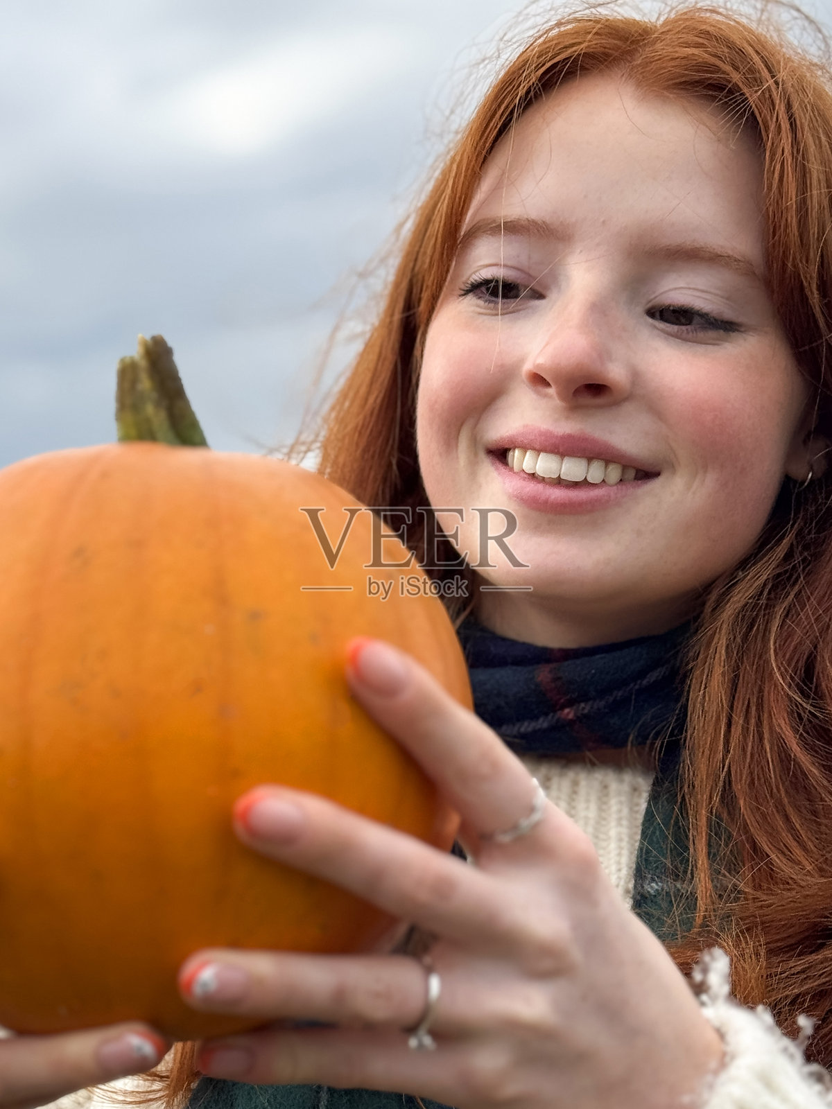 特写镜头，迷人的红发年轻女性穿着阿兰毛衣和格子围巾，手持橙色南瓜，展示修饰过的彩绘指甲，万圣节设计的橙色法式指尖美甲上有白色鬼魂，阴郁的灰色天空，焦点在前景。照片摄影图片