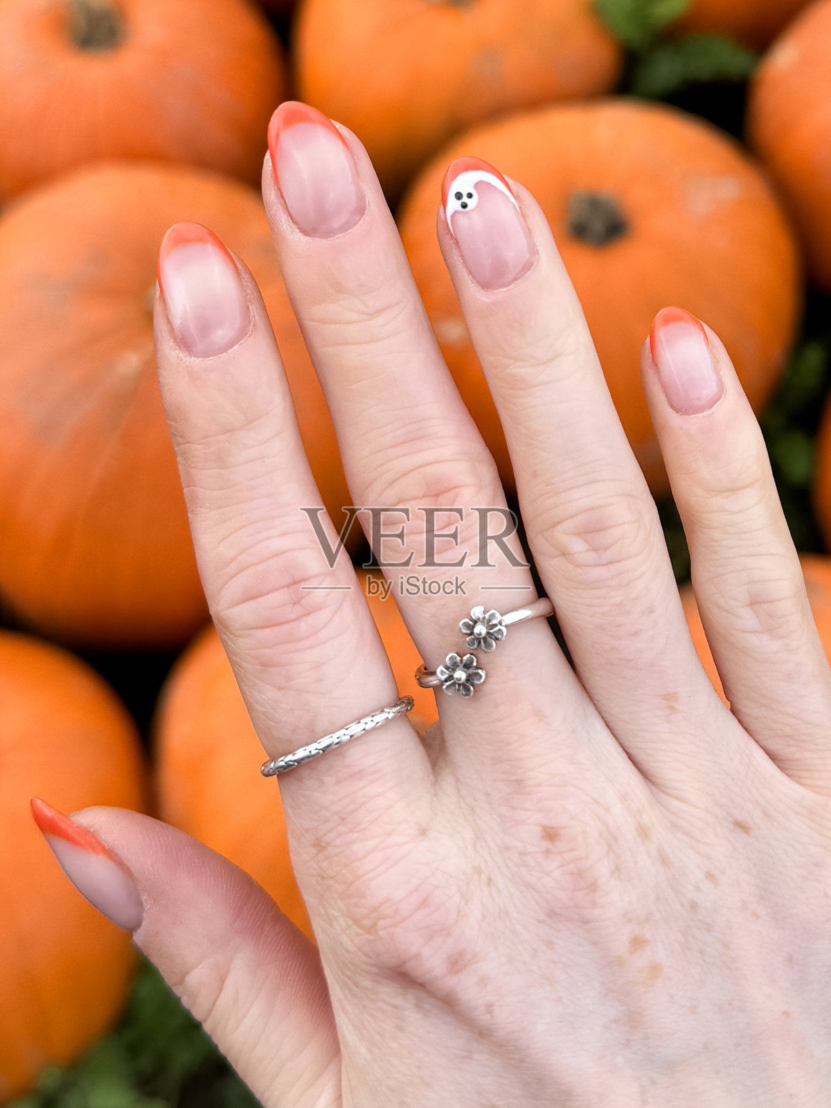 特写镜头，无法辨认的女性佩戴银色戒指，手部展示修饰过的彩绘指甲，万圣节设计的橙色法式指尖美甲，带有白色鬼魂，户外环境，背景为一组橙色南瓜，俯视角度。照片摄影图片