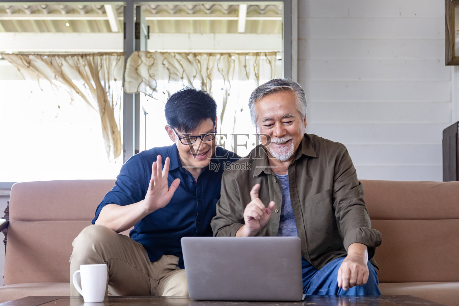 一位年长的亚洲老人和他的儿子通过电脑视频通话向家人问好，他们在舒适的退休之家，旁边留有空白空间。照片摄影图片