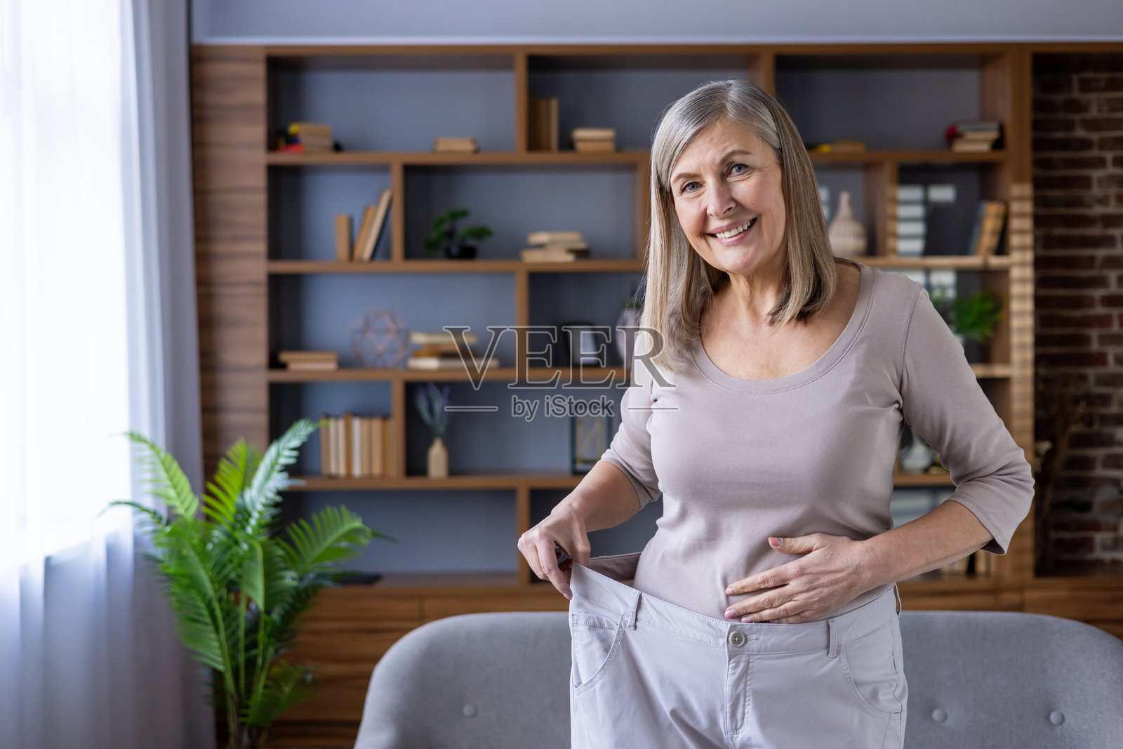 微笑的成熟的女性在室内轻松的环境中展示减肥成功，手中拿着一条大裤子。照片摄影图片