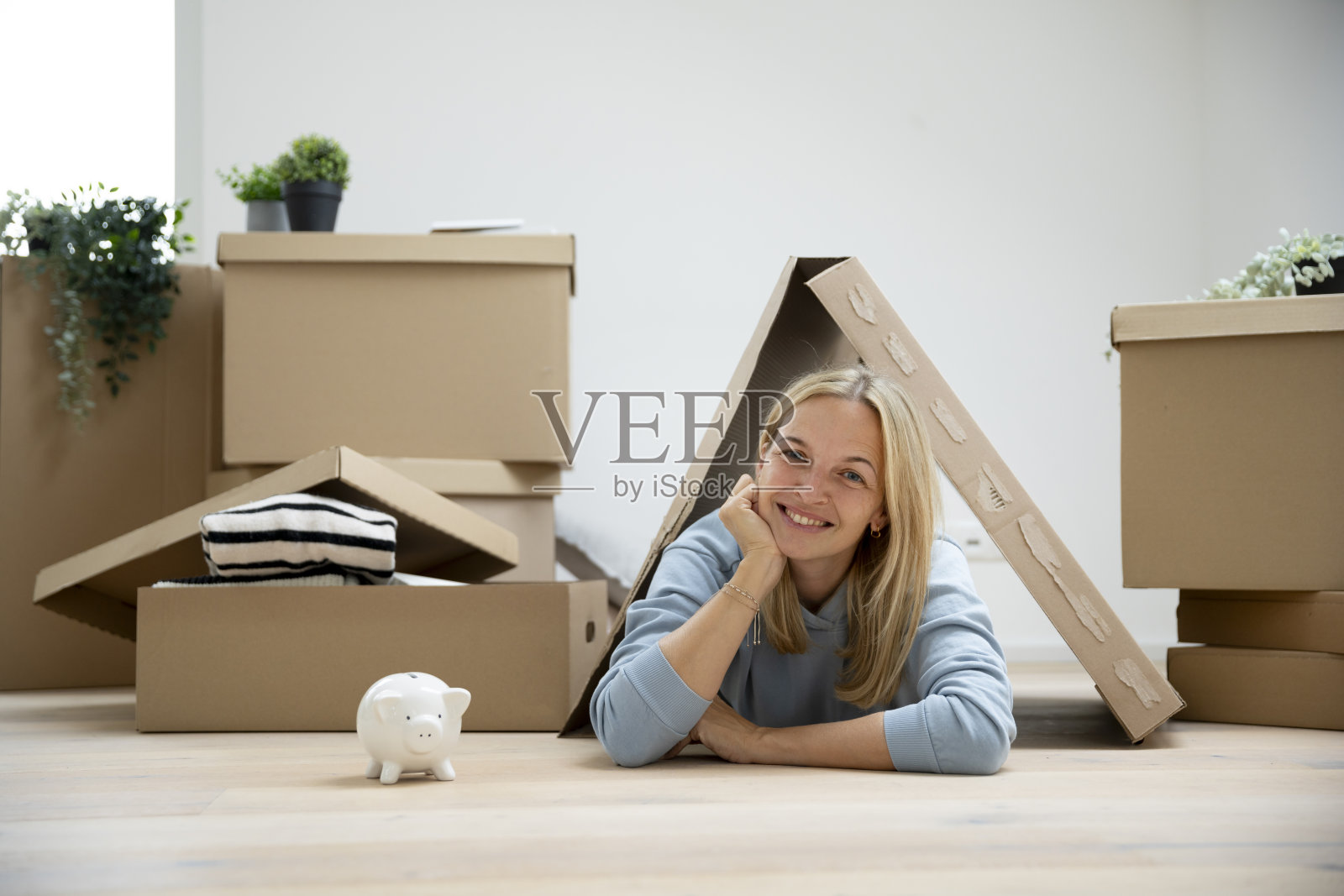 年轻的金发女性在她的新公寓里，手里拿着储蓄罐，周围有很多搬家的箱子。照片摄影图片