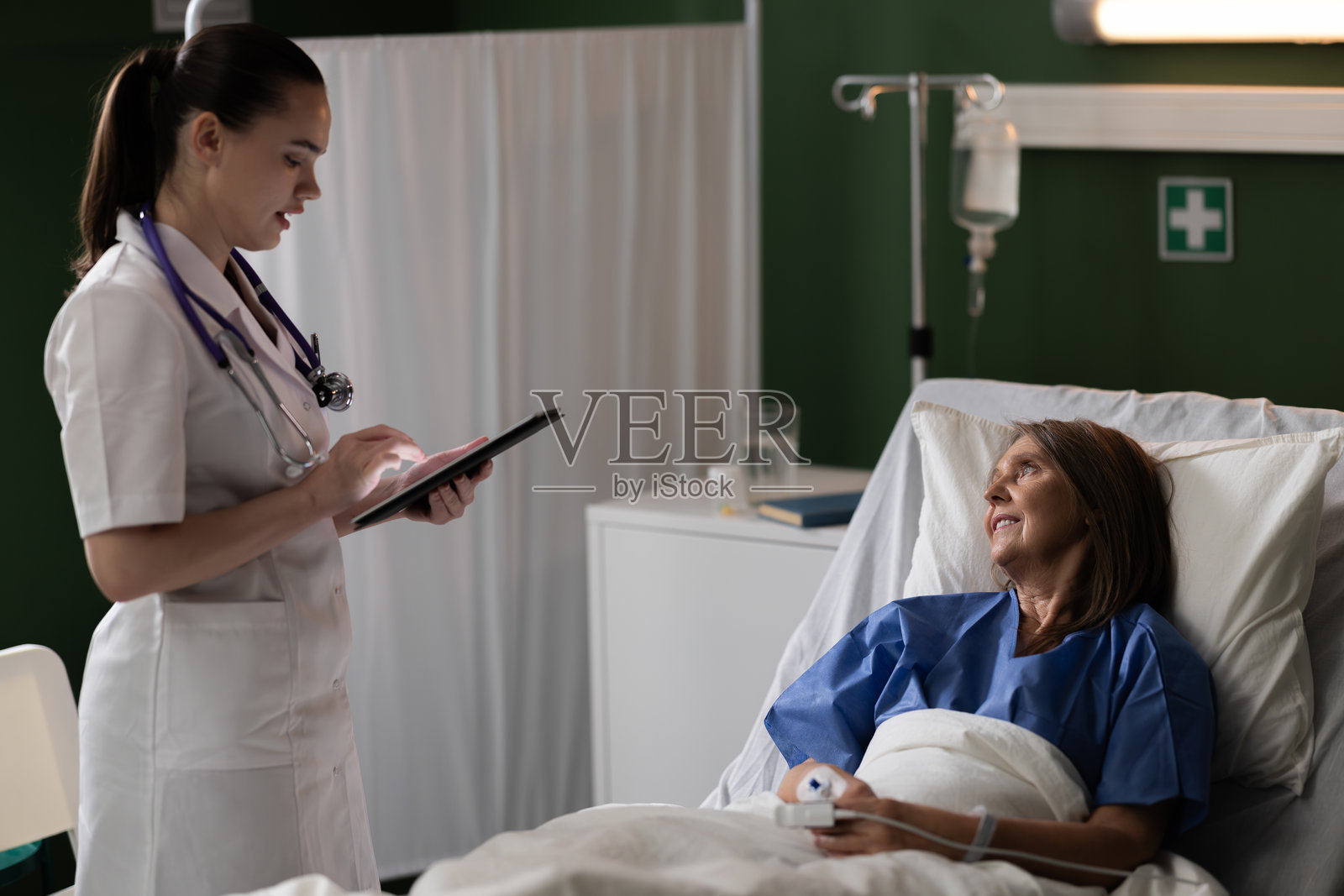 年轻医生在医院病房探望一位老年女性，手中拿着平板电脑询问她的健康状况。照片摄影图片