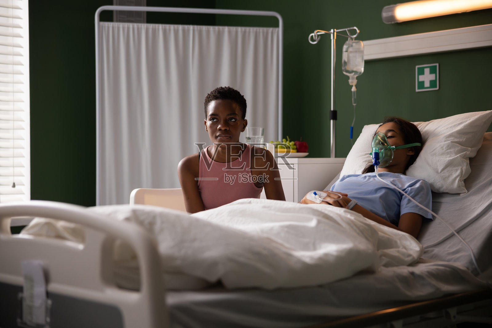 悲伤的非洲母亲坐在医院病房里，陪伴着躺在床上、戴着氧气面罩的女儿。照片摄影图片