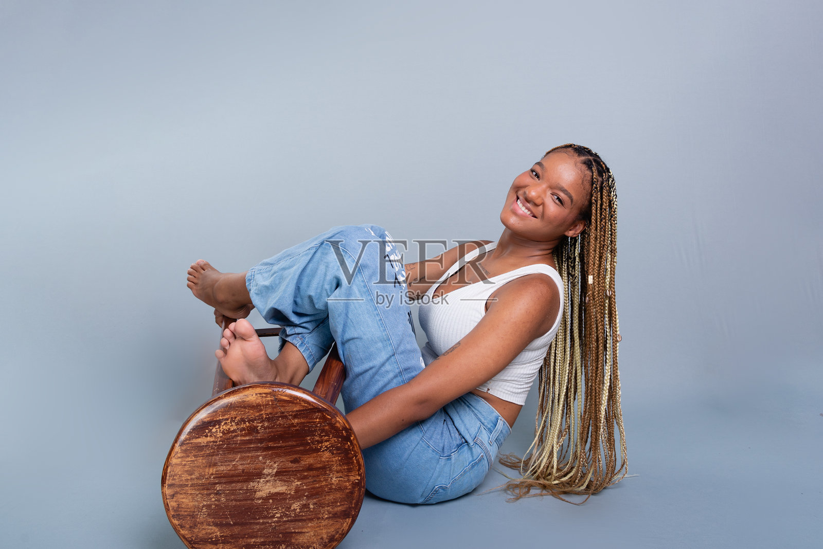 一位微笑的棕发女性，头发编成辫子，坐在工作室的地板上，靠在一张木凳子上。照片摄影图片