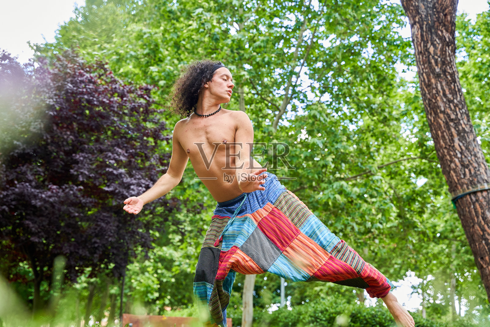 在郁郁葱葱的绿意中，一位赤裸上身、长发卷曲的男子穿着宽松裤子，优雅地舞动，展现出自由与表达的当代舞蹈。照片摄影图片