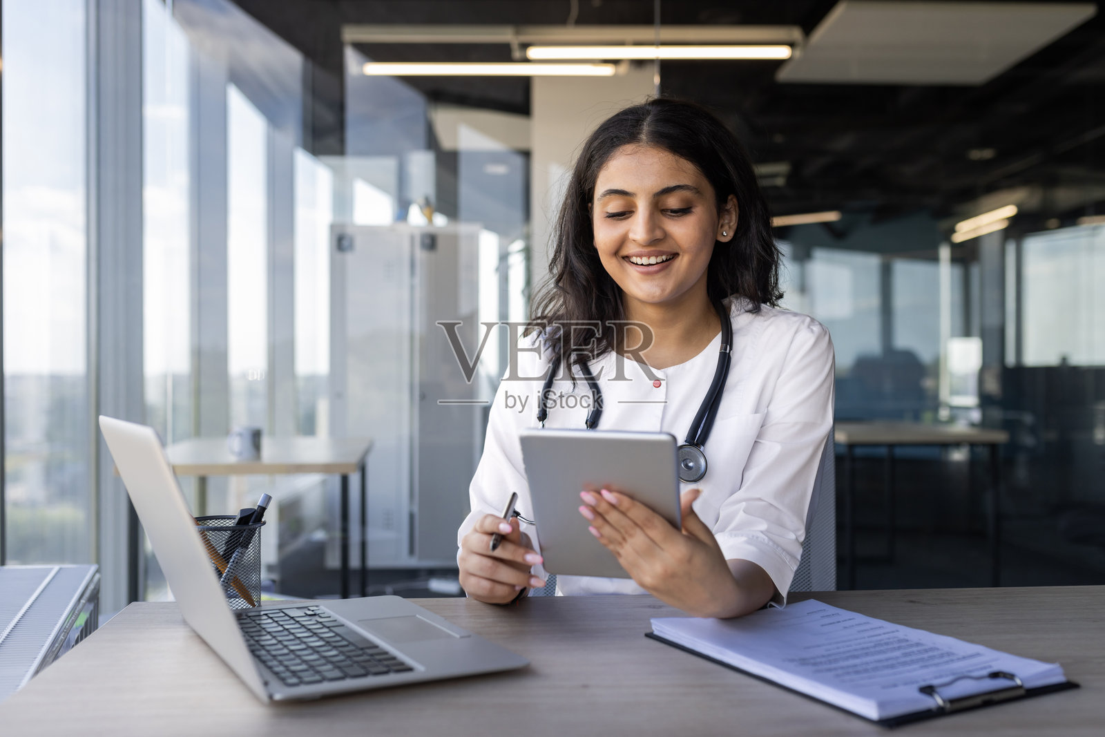 医生在办公室环境中微笑着使用平板电脑，桌上有笔记本电脑和文件。照片摄影图片