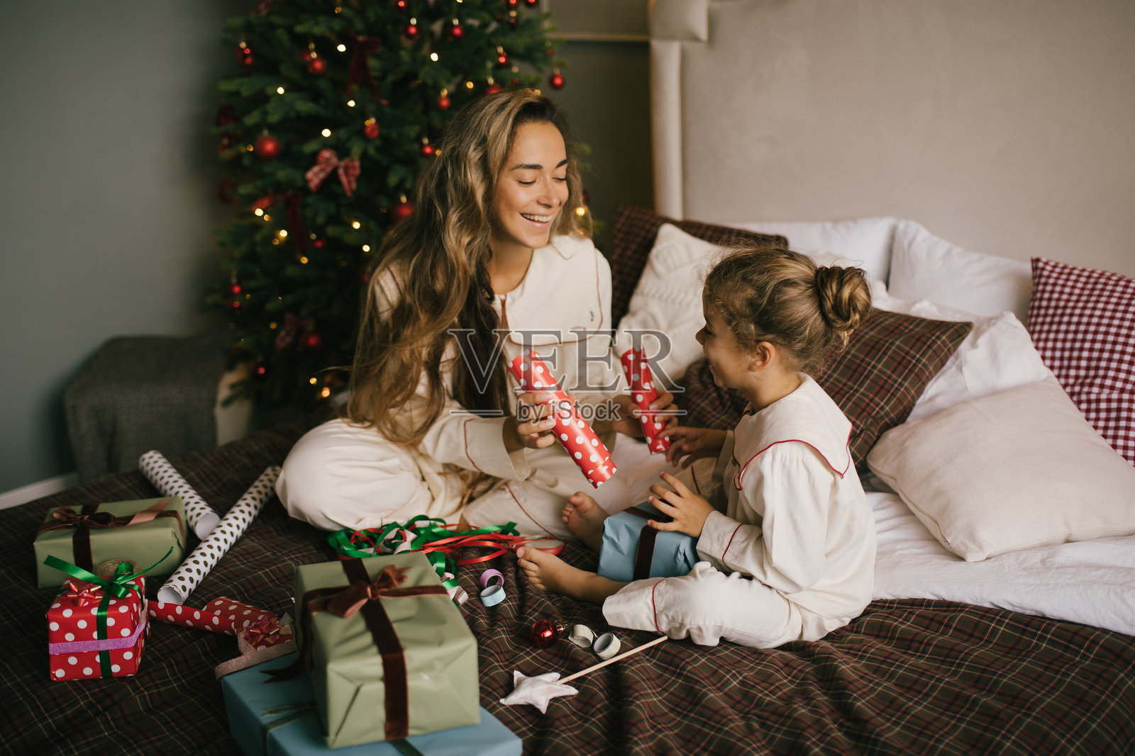 年轻漂亮的母亲和她的女儿在温馨装饰的卧室里打包和包装礼物，旁边有一棵圣诞树。照片摄影图片
