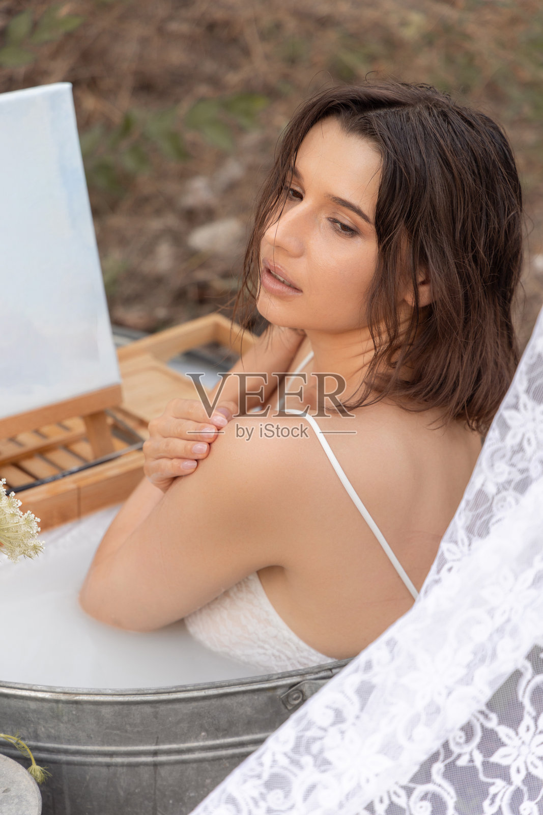 迷人的女性在户外浴缸中。女孩坐在牛奶水中，穿着裙子，在画架上作画。女性在花园中放松，靠在复古的旧门和墙上。树上挂着窗帘。照片摄影图片