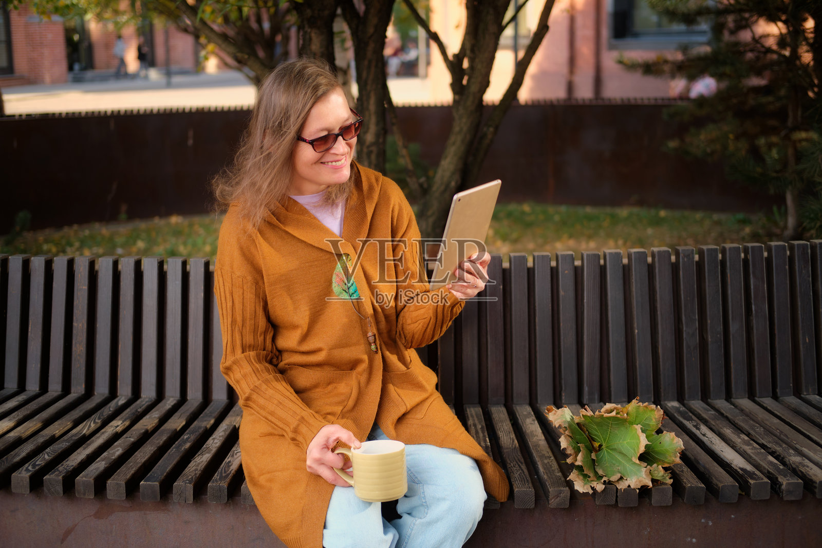 微笑的女性穿着秋季服装，手持平板电脑和咖啡杯，坐在公园长椅上，周围是落叶。户外生活方式概念。50岁女性。照片摄影图片