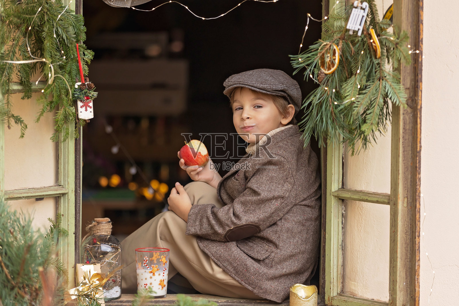 美丽的金发男孩坐在装饰有圣诞节装饰的窗边，喝着牛奶，吃着苹果，等待圣诞老人，圣诞节概念照片摄影图片