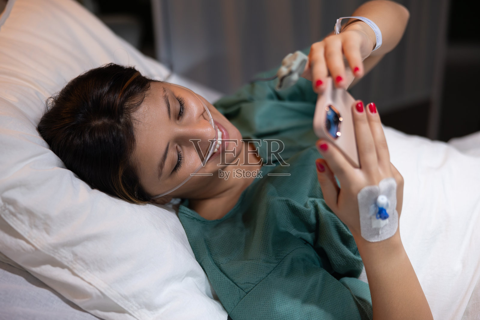 年轻女孩在氧气管中躺在病房里接受治疗，手中拿着手机，查看照片。照片摄影图片