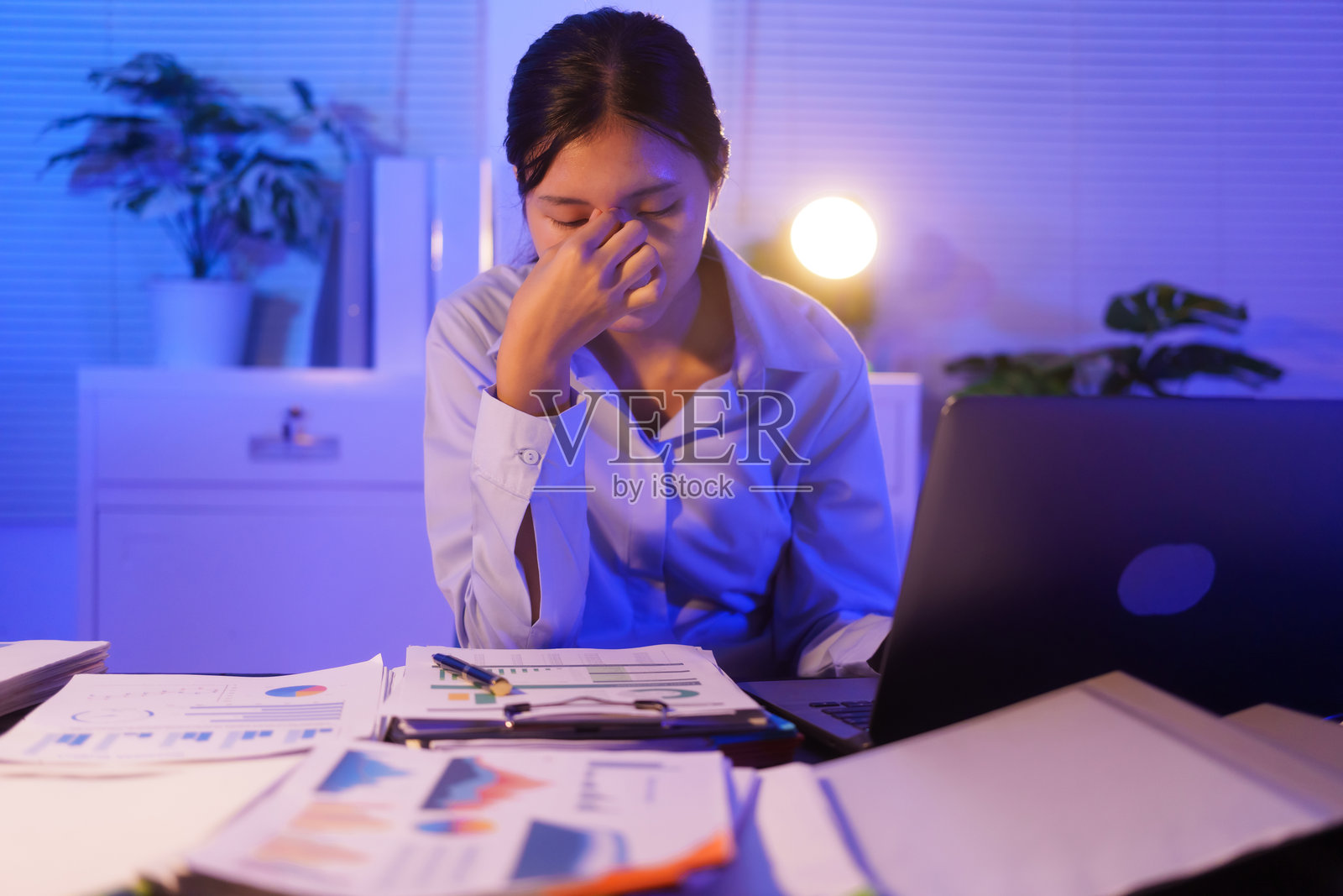 一位年轻女性在办公室的桌子上加班，使用笔记本电脑审阅文件。她感到压力、疲惫，因久坐而头痛、身体酸痛，导致办公室综合症。照片摄影图片