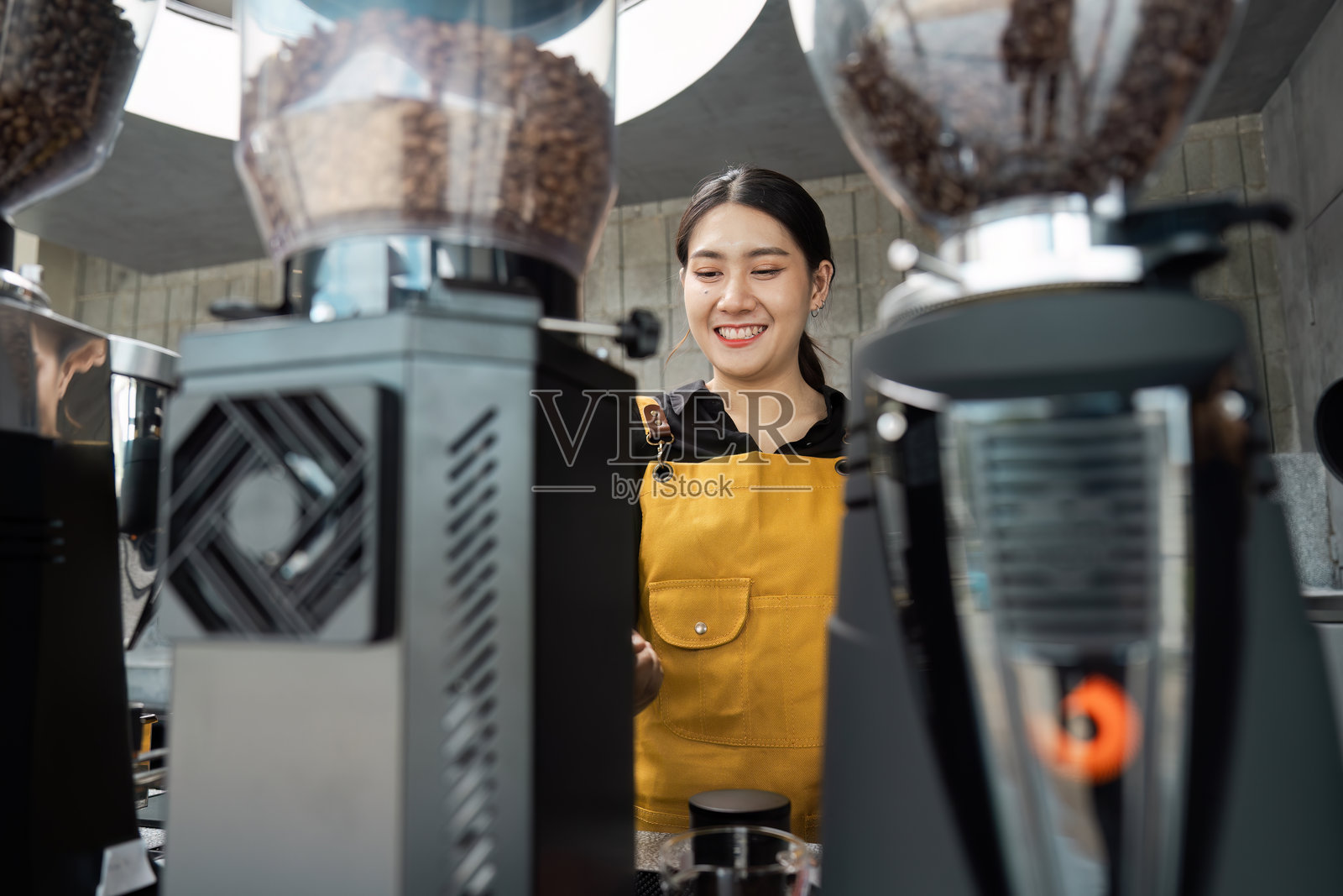 穿着黄色围裙的咖啡师在现代咖啡馆中微笑着准备咖啡，周围是专业设备。照片摄影图片